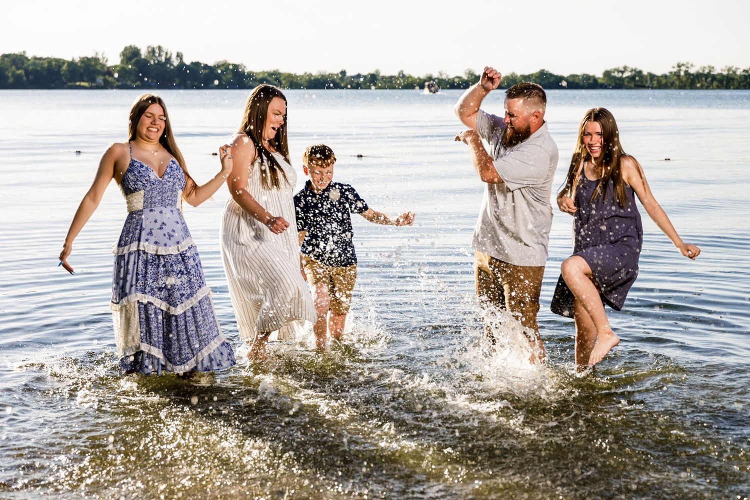 Family of five joyfully splashing through shallow lake water on a sunny day, wearing summer dresses and casual clothes.