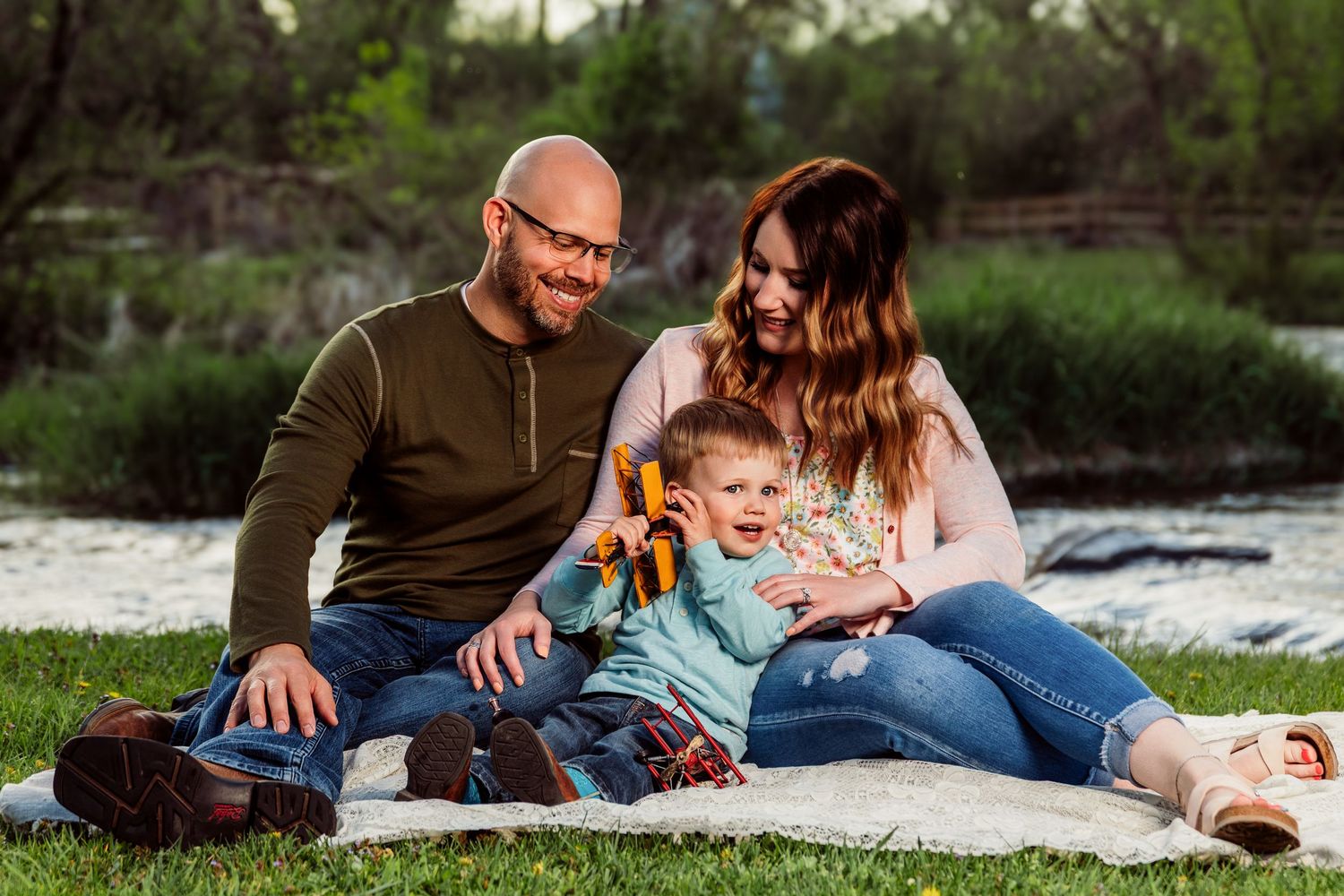 Happy family with toddler sitting on blanket by river, smiling outdoors in casual clothes during golden hour.