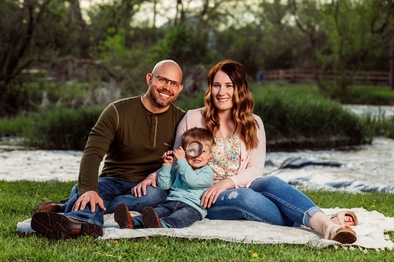 Happy family of three sitting on a blanket by a pond, smiling outdoors in a lush green park setting.
