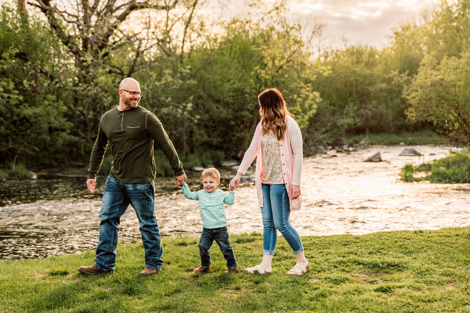 Family of three holding hands and walking by a sunlit river surrounded by lush green trees and grass.