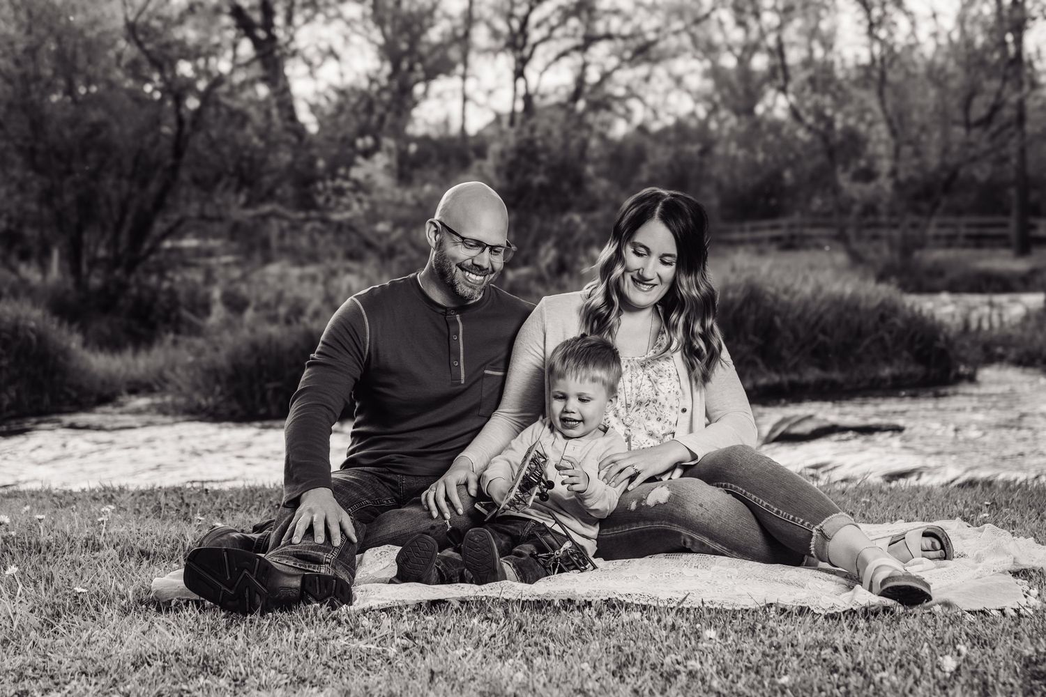 Black and white family photo of parents and toddler sitting on blanket outdoors near trees.