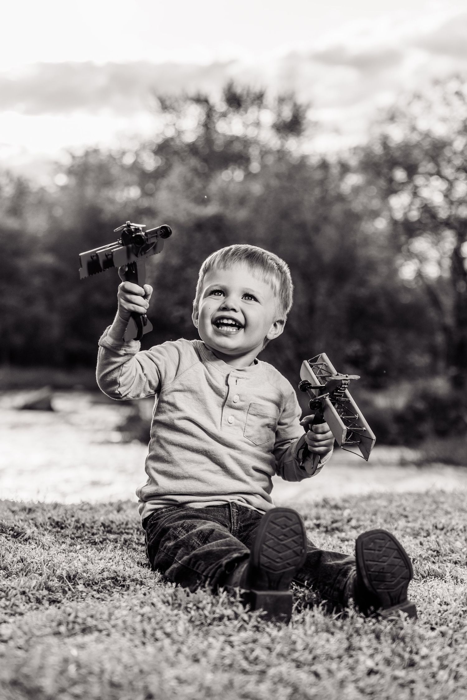Black and white photo of a happy toddler boy sitting on grass outdoors, holding two toy airplanes while smiling.