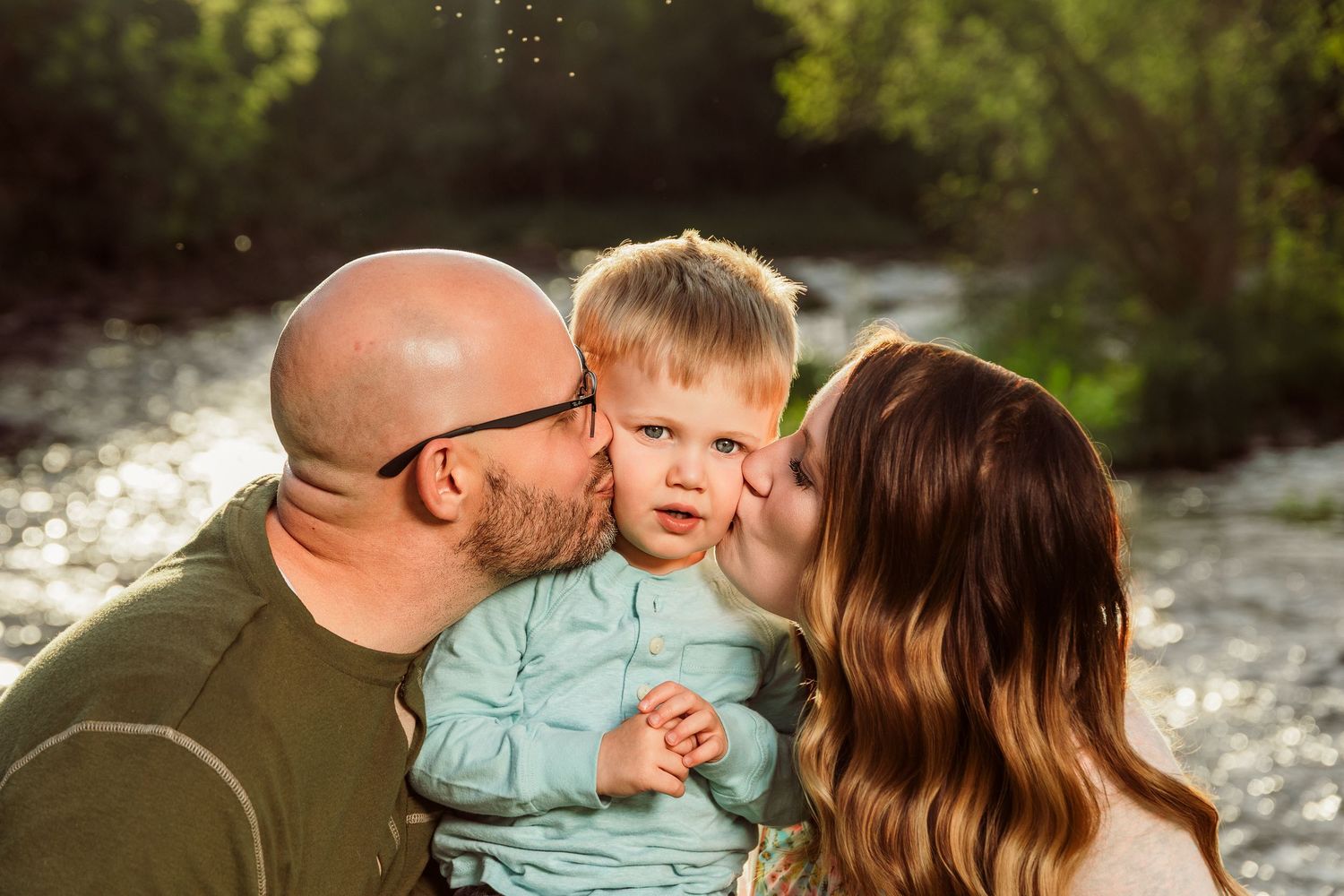 Parents kissing their toddler boy on cheeks outdoors near a sunlit stream in a warm family portrait.