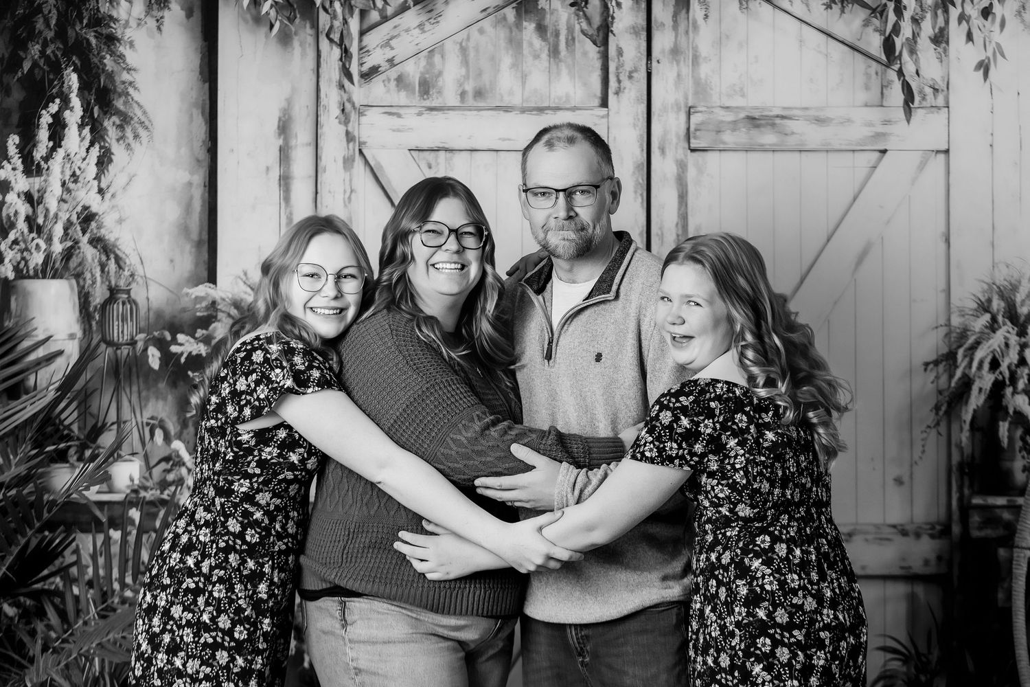 Black and white family portrait of four people hugging and smiling in front of rustic barn doors with greenery.