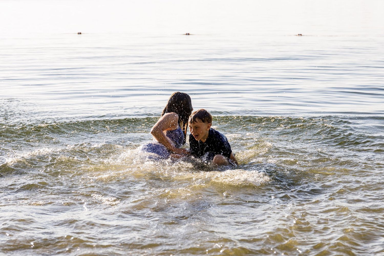 Two children sitting together in shallow lake water, smiling and enjoying a sunny summer day.