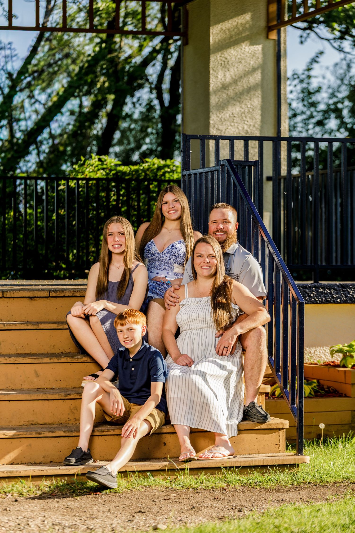 Happy family of five posing on sunny outdoor steps, smiling together in casual summer outfits.