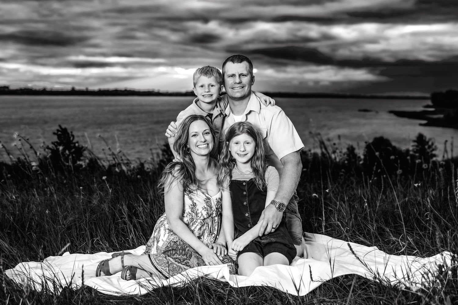A loving family bonding moment captured in black and white against a dramatic cloudy sky at sunset.