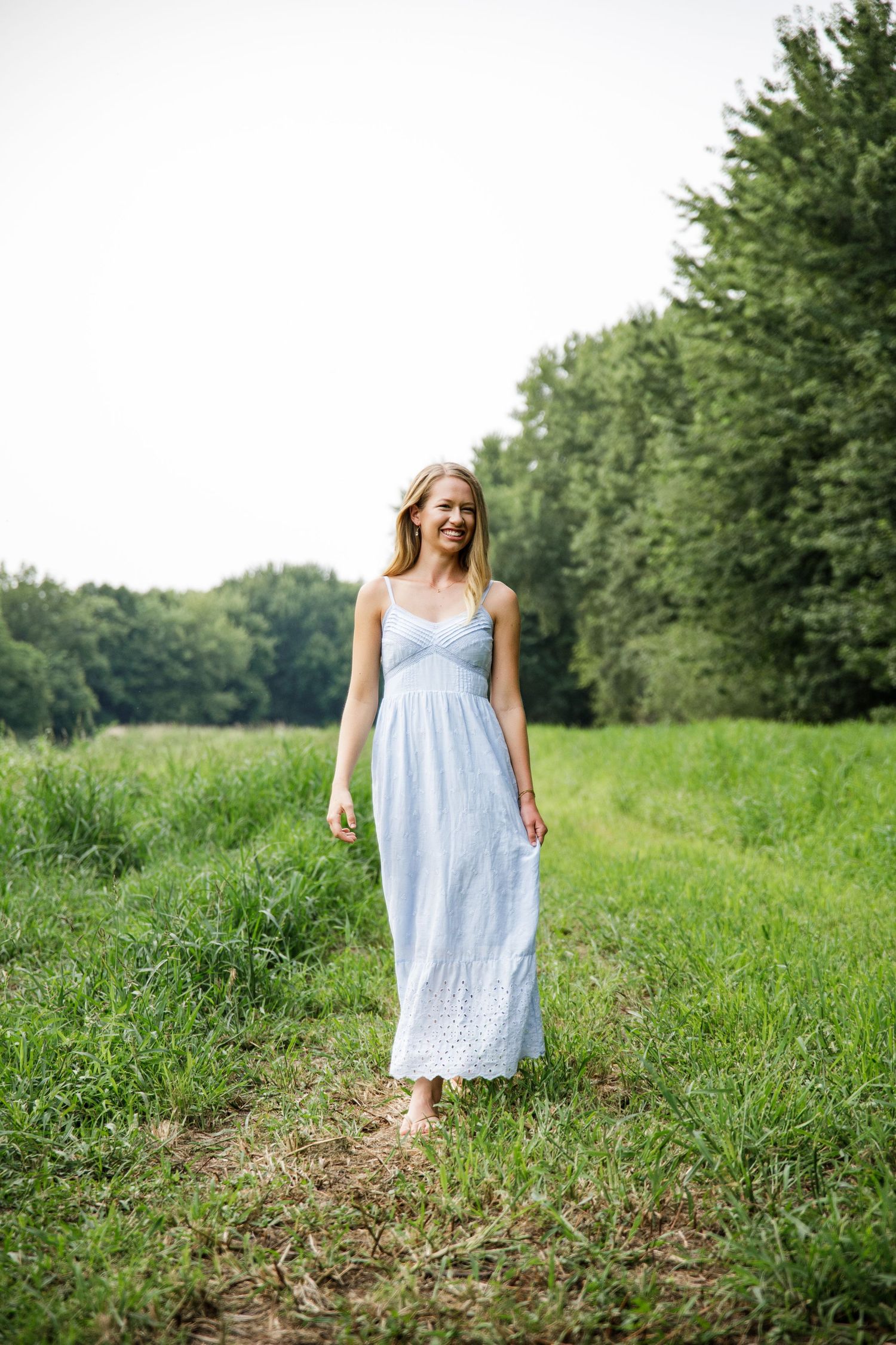 Young woman in light blue sundress walking barefoot through green meadow with trees in background.