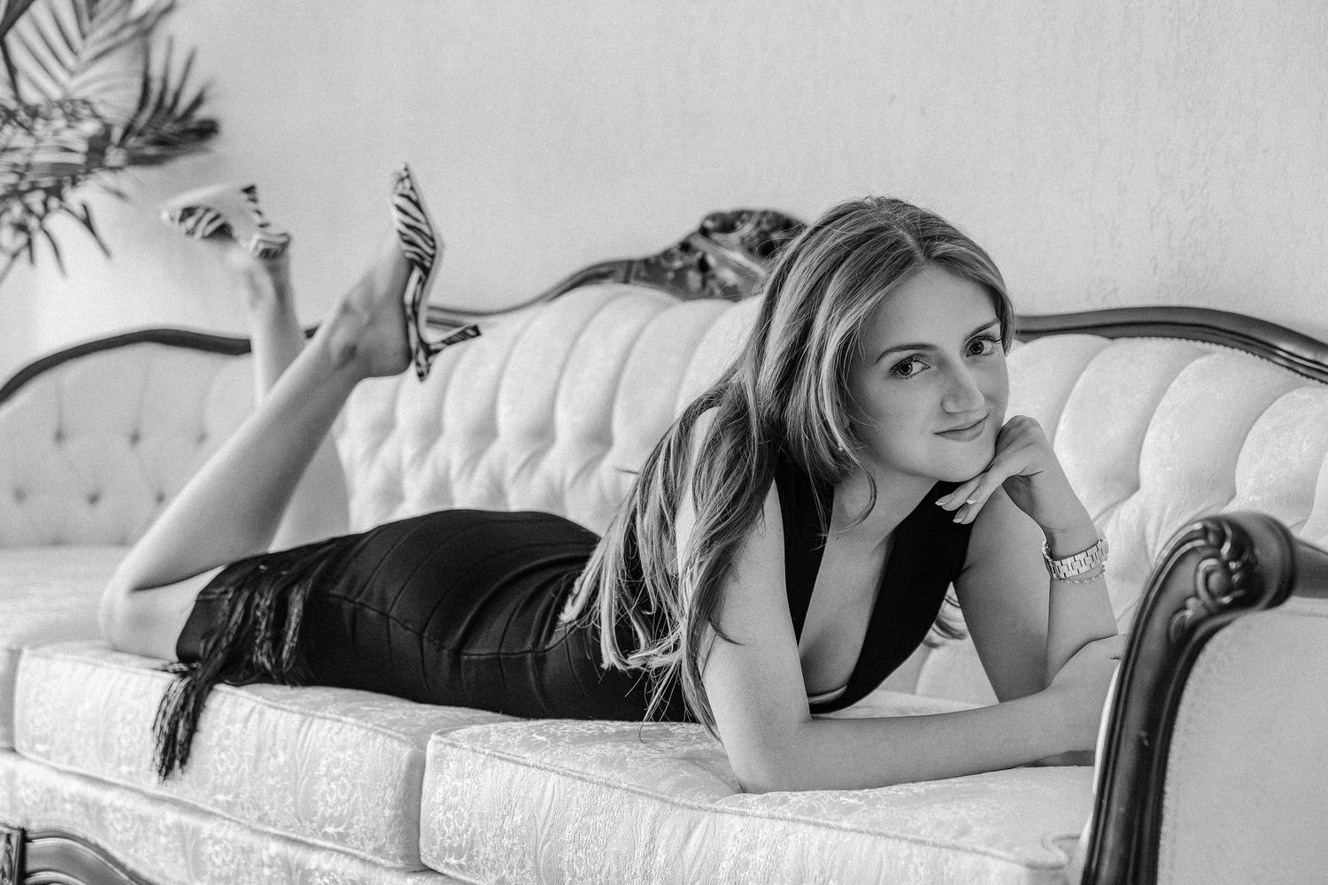 Young woman in black dress poses elegantly on a vintage tufted sofa in a black and white portrait photo.
