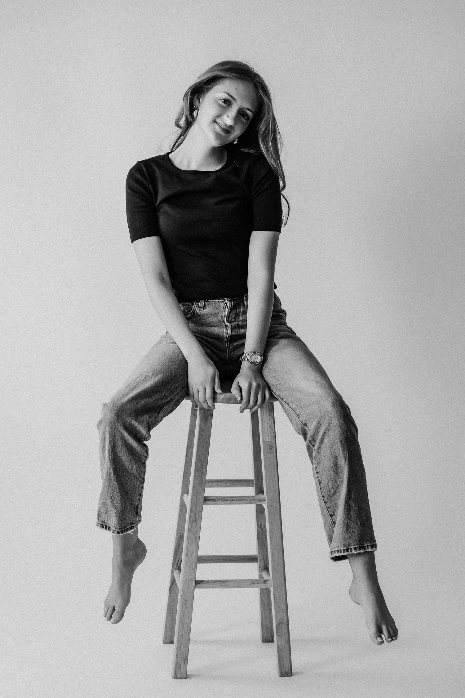 Black and white full-body portrait of smiling young woman sitting on wooden stool in black tee and jeans, barefoot.