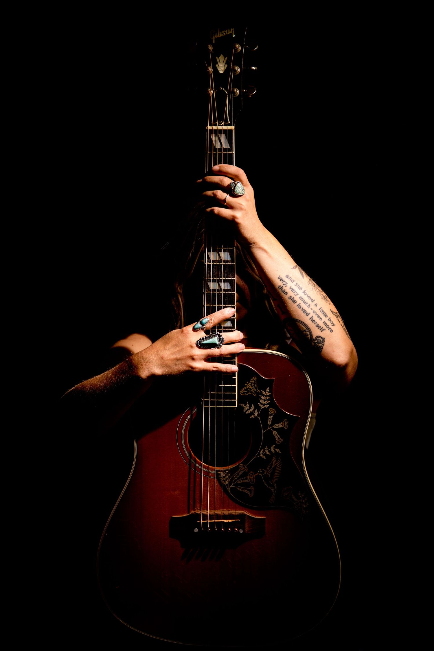 Tattooed hands holding a dark red acoustic guitar against a black background in dramatic low-key lighting.