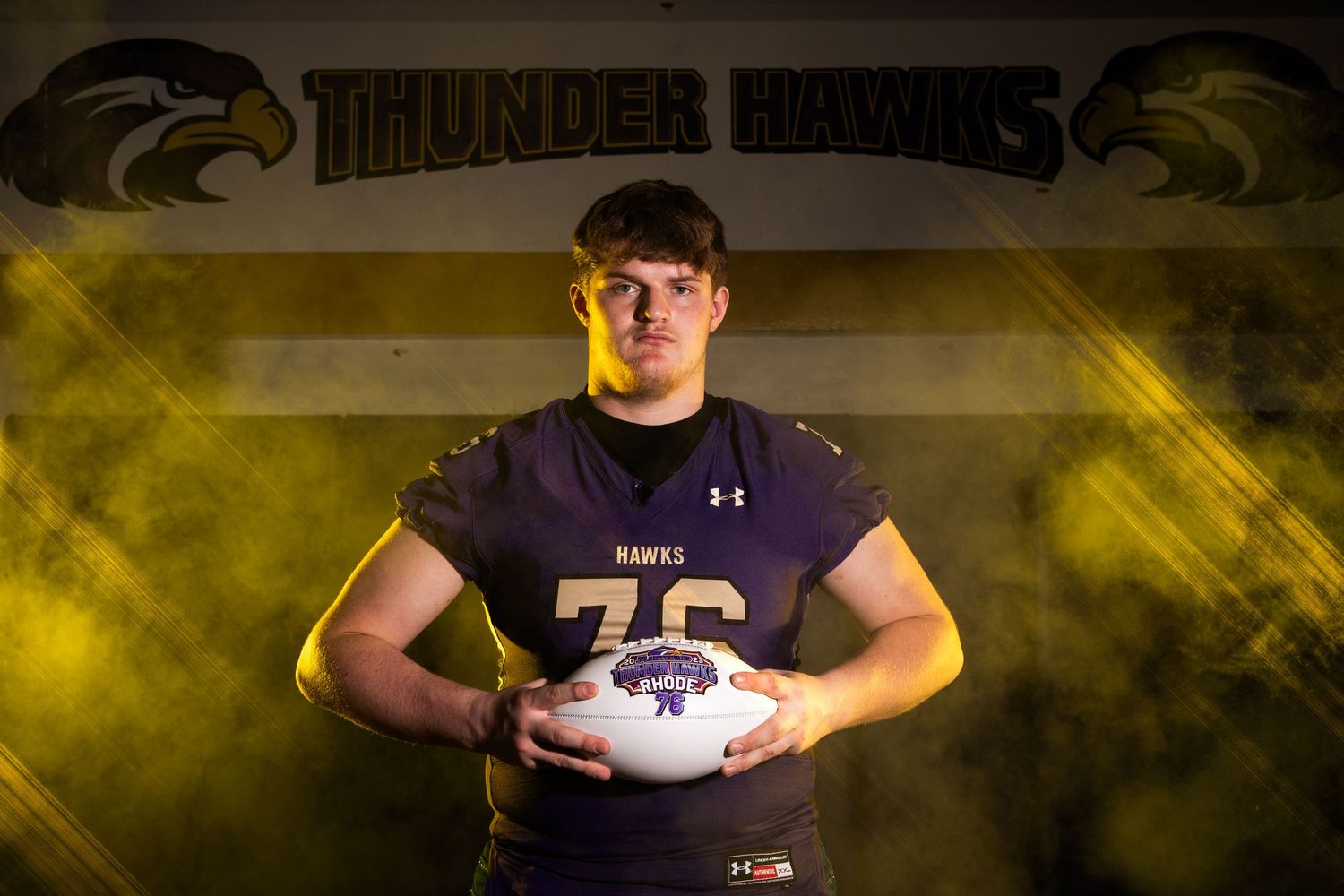 High school football player in uniform holds white football while posing against dramatic yellow backdrop.