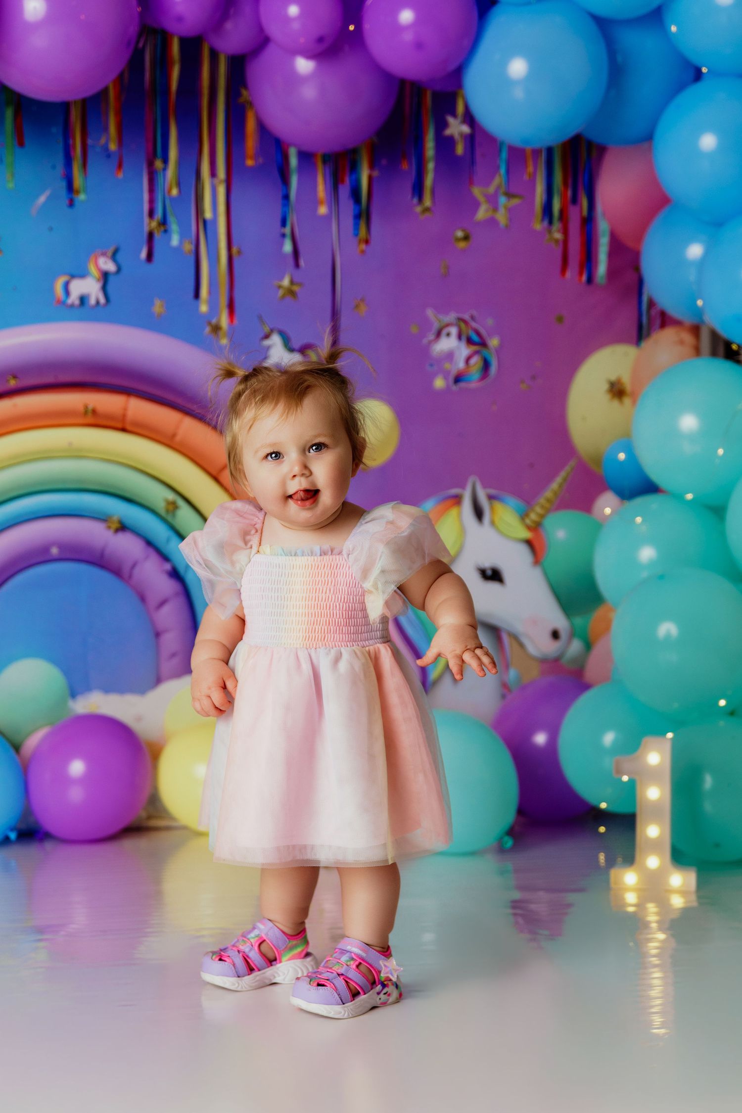 Toddler girl in pink dress at unicorn rainbow balloon first birthday party celebration.