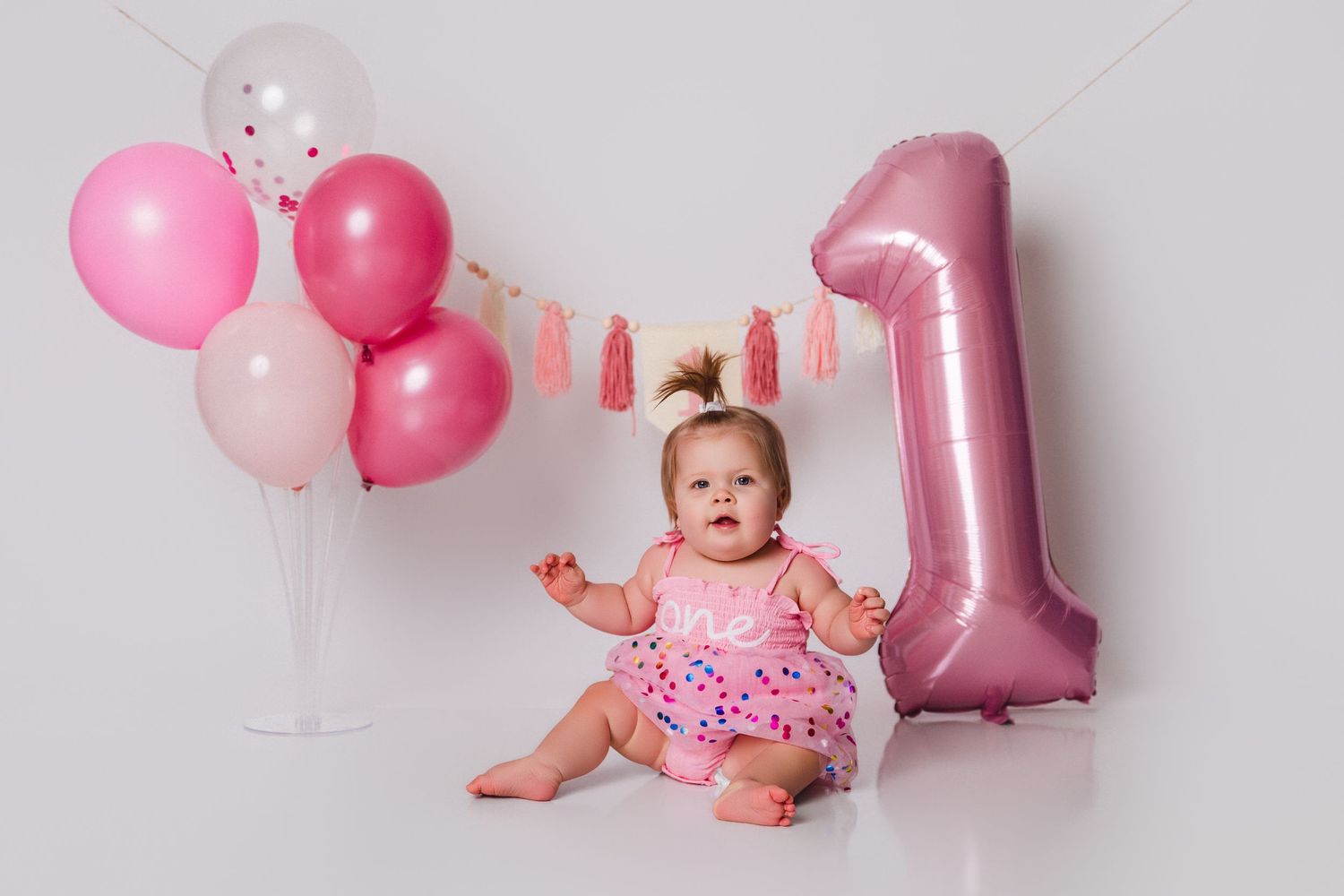 Baby sitting among pink balloons and a number one balloon for first birthday photos.