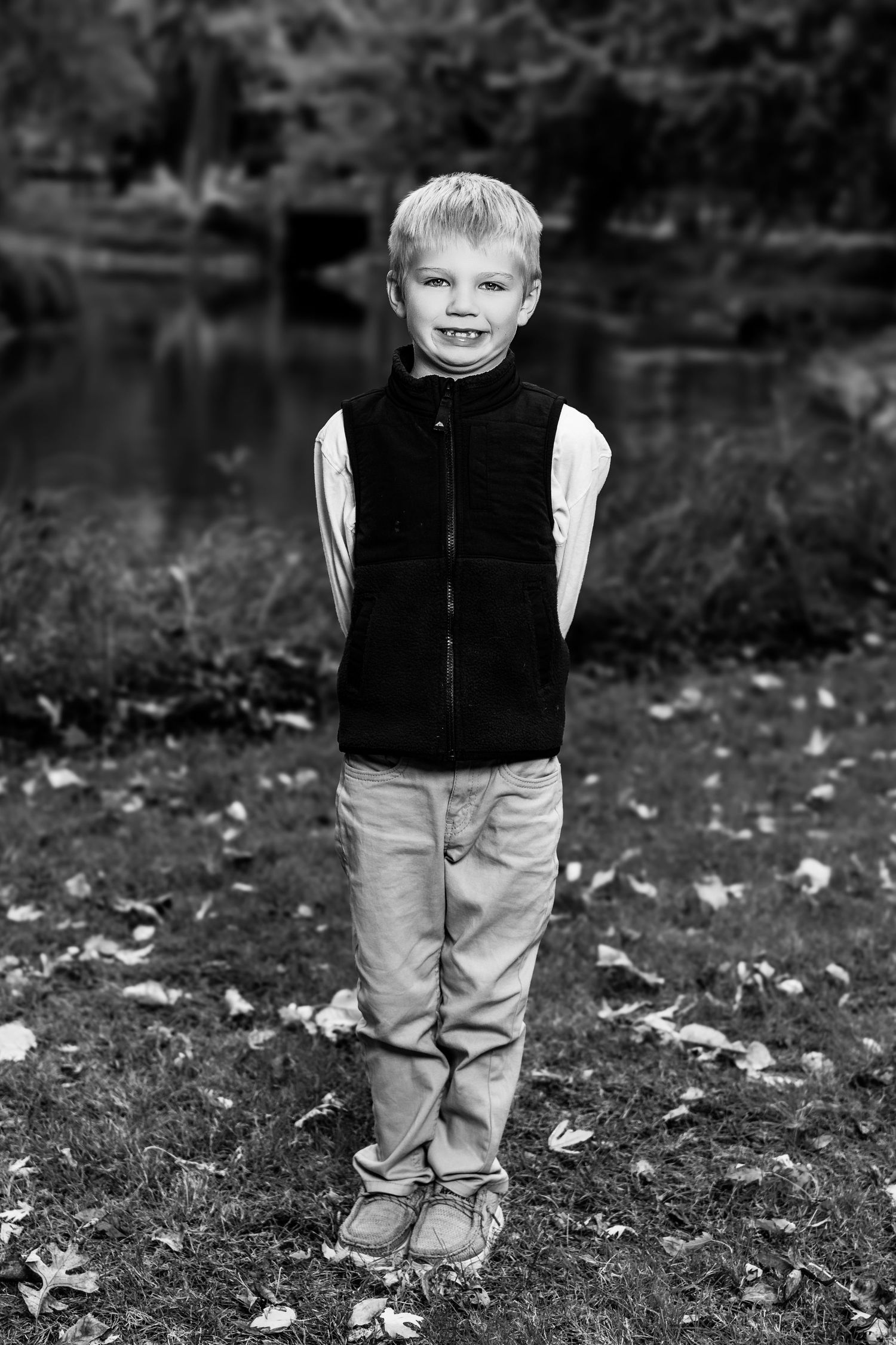 Black and white portrait of a small child wearing a black vest and khaki pants standing on grass with fallen leaves.