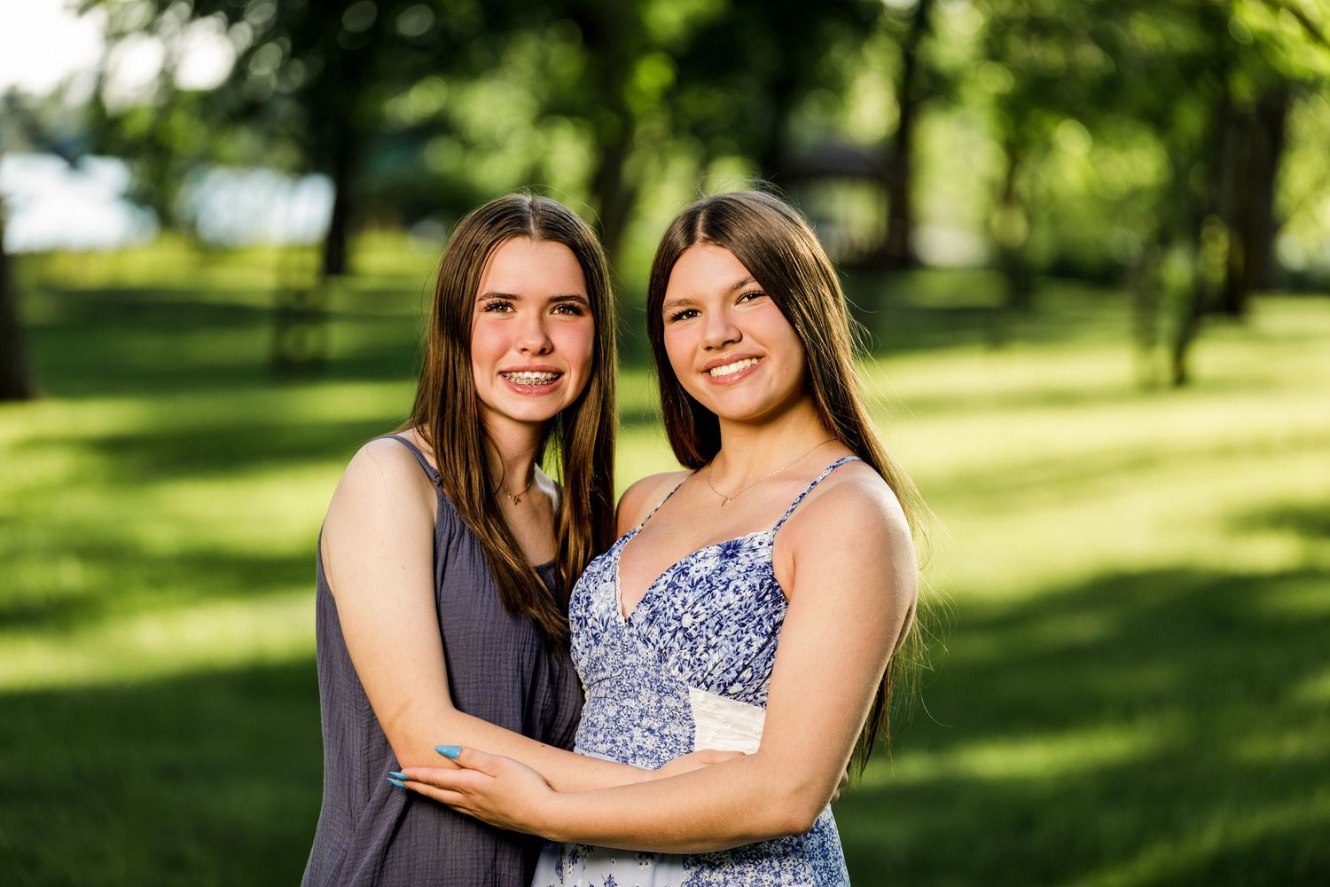 Two teenage girls embracing and smiling together in a sunny green outdoor park setting.