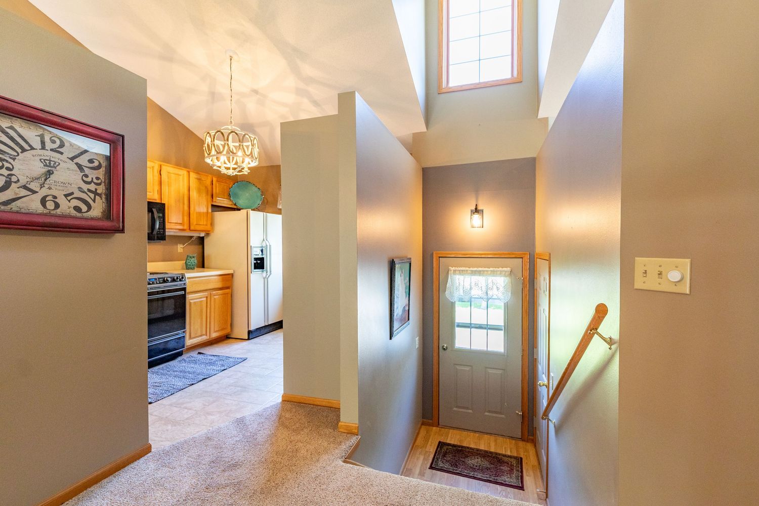 Bright entryway with vaulted ceiling, chandelier, front door with window, and open kitchen with wood cabinets and white appliances.
