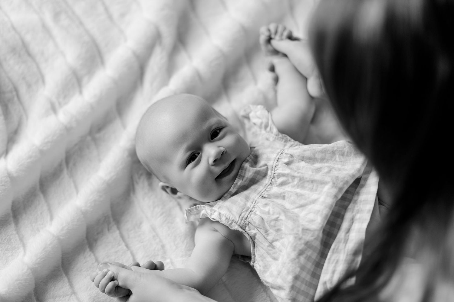 Black and white close-up of a smiling baby lying on a blanket looking up at their mother.