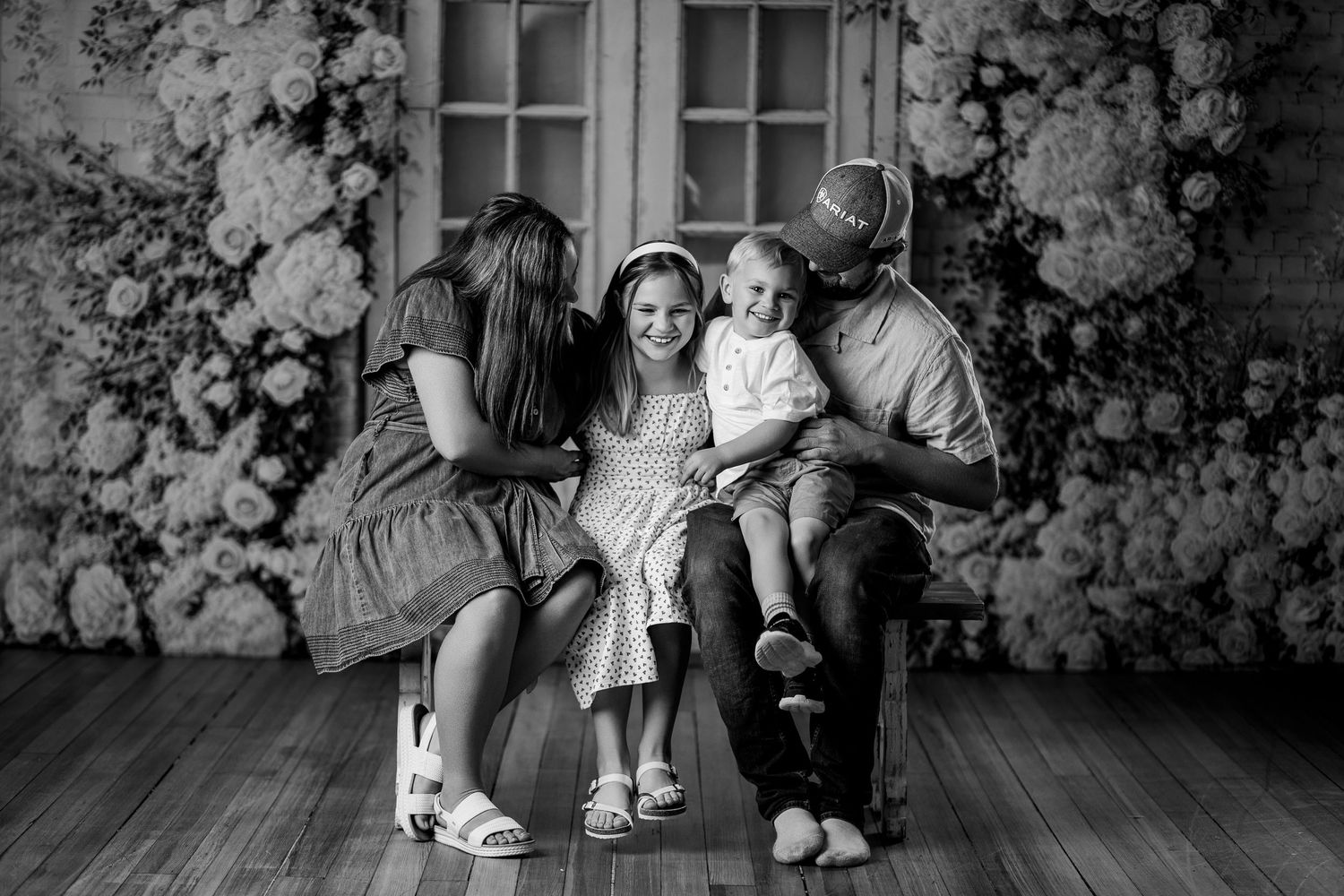 Black and white family portrait with parents and two young children sitting together in front of a floral backdrop.