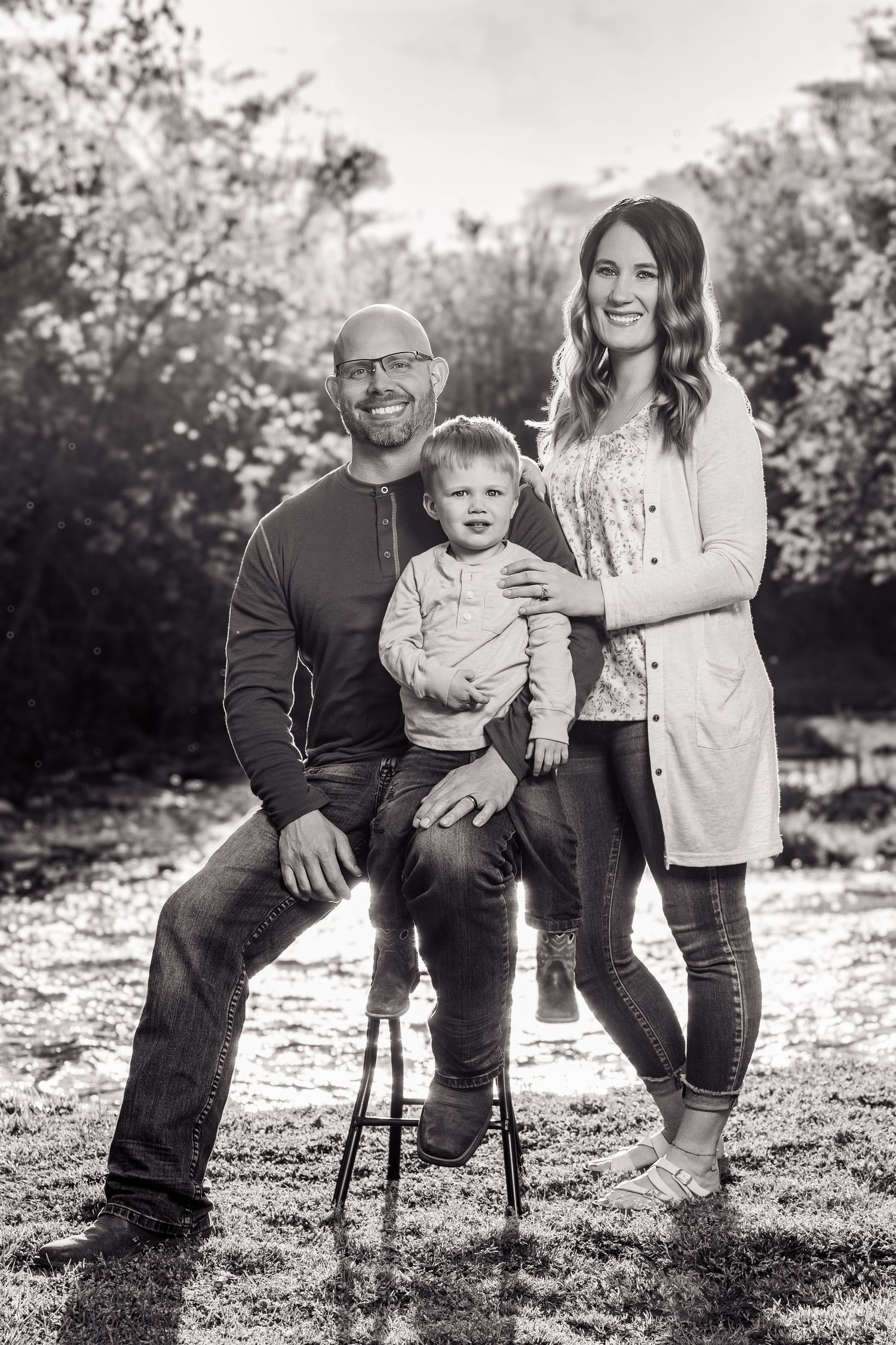 Black and white family portrait of a bald man, woman, and toddler posing outdoors in a park setting.