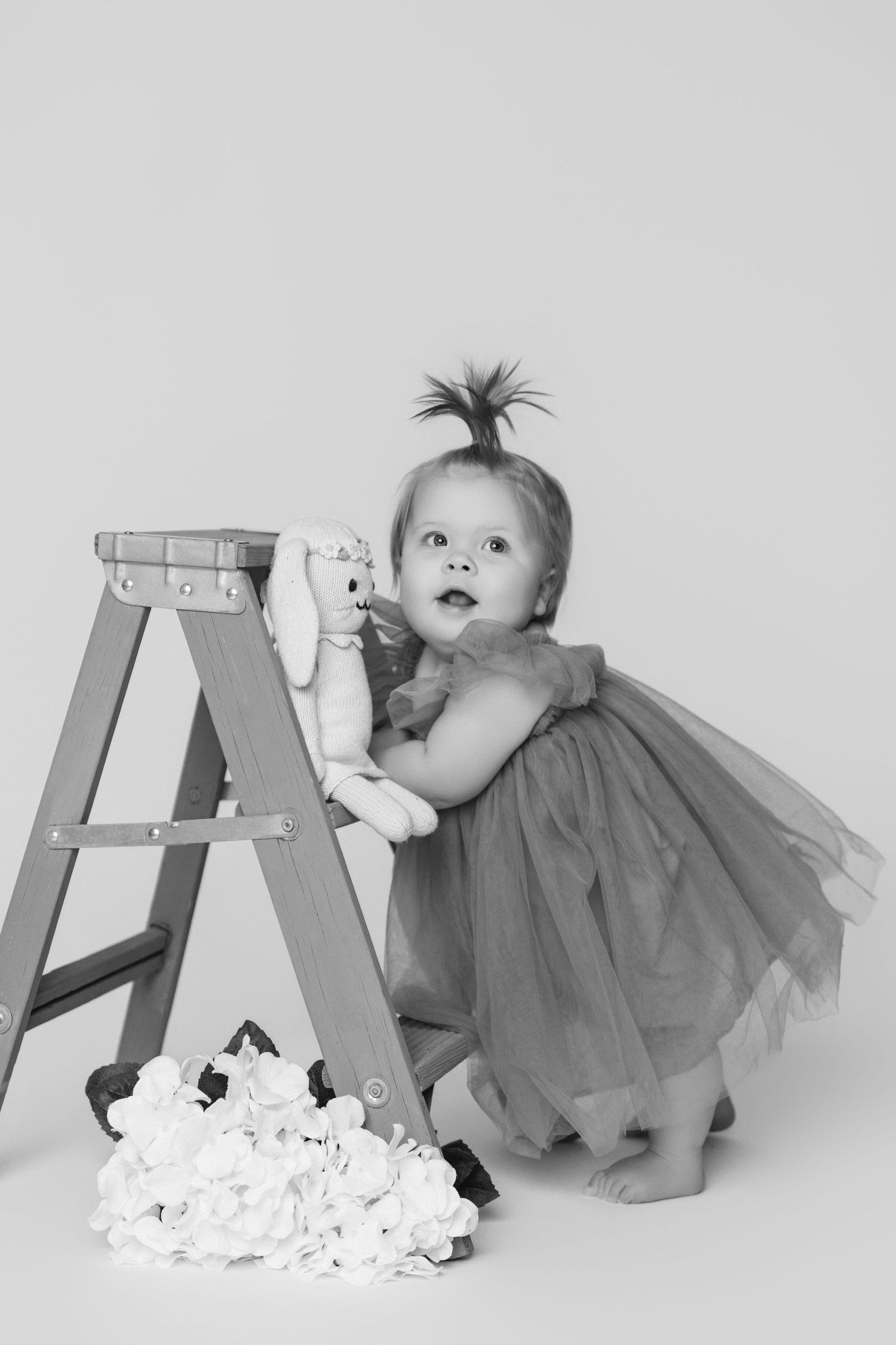 Baby in grey dress crawling near wooden ladder with scattered paper flowers.