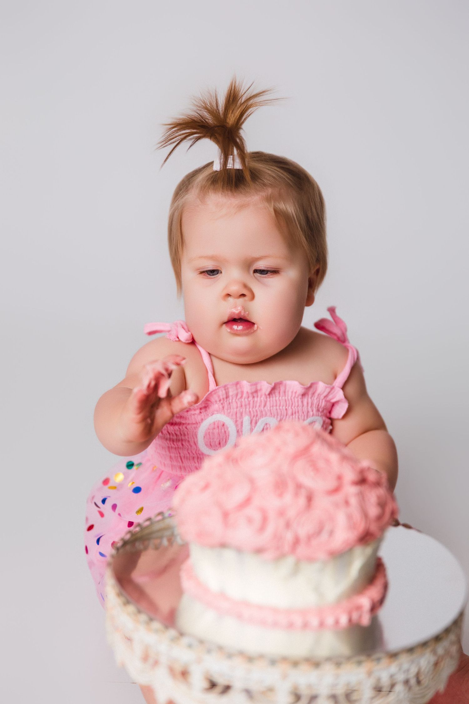 Baby in pink outfit enjoying first birthday cake smash photo session.