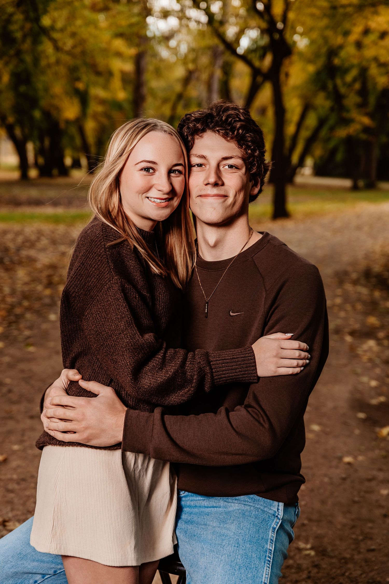 A couple embraces while wearing brown sweaters during an autumn photoshoot with golden trees in the background.