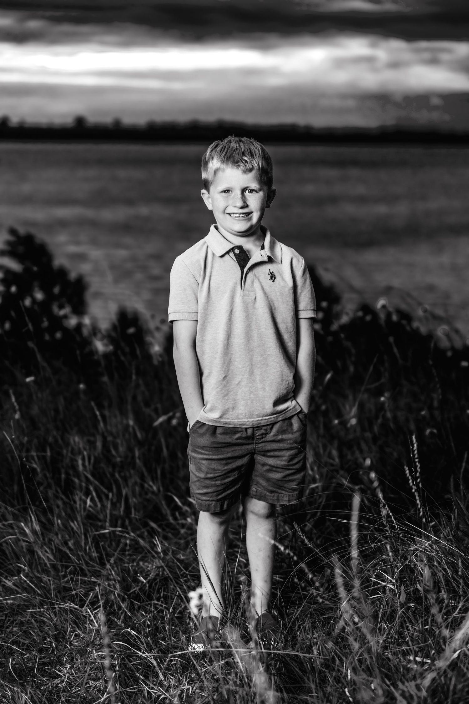 Black and white portrait of a young child standing in tall grass near a lake at sunset, wearing a polo shirt and shorts.