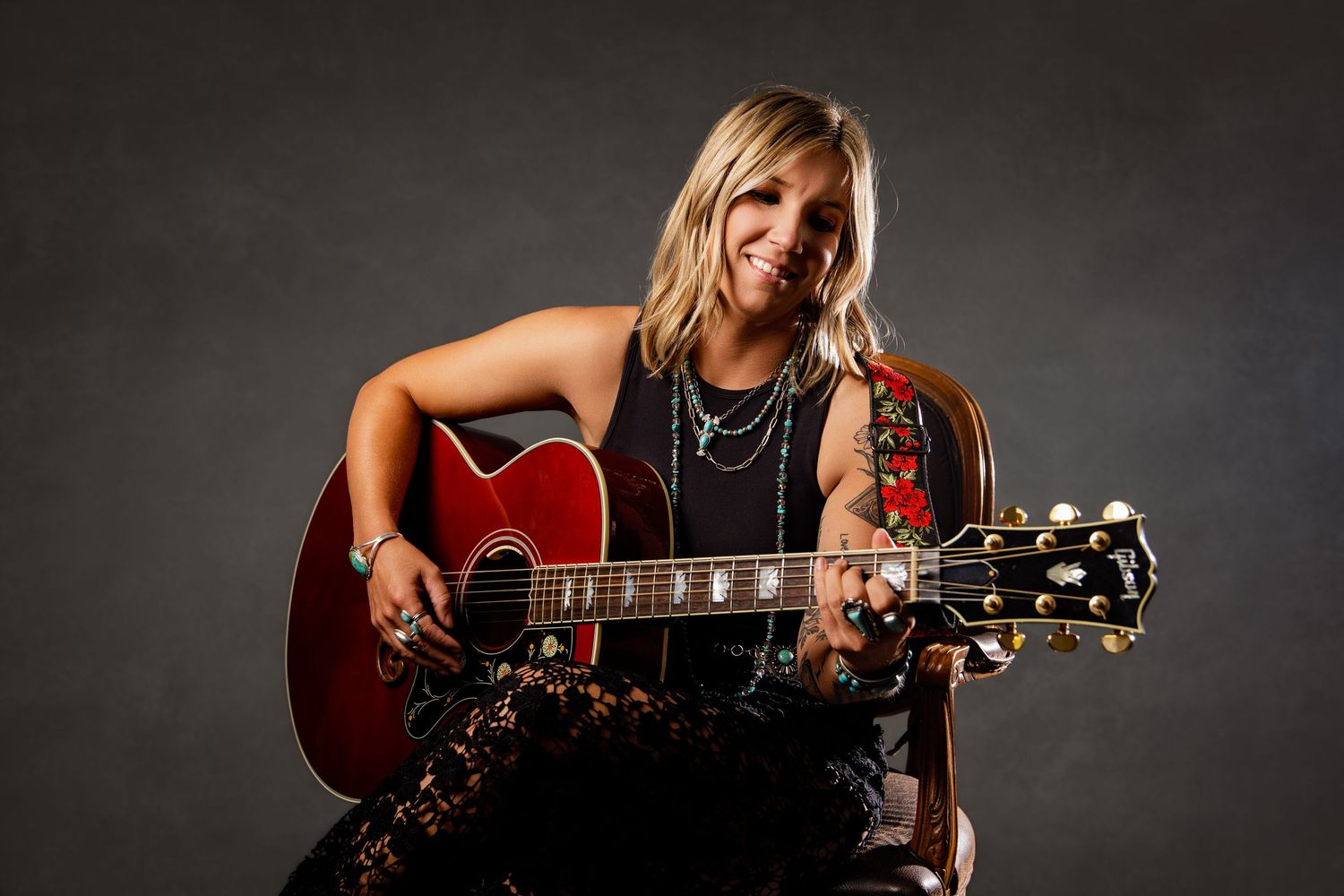 Smiling blonde woman playing a red acoustic guitar, wearing a black outfit with layered necklaces, posed against a gray background.