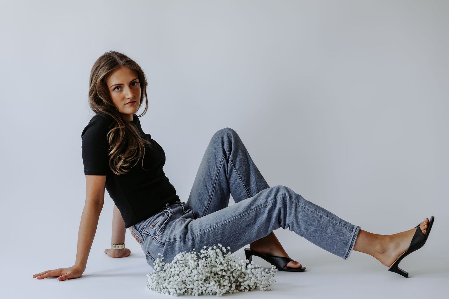 Woman in black top and jeans sitting on white floor with baby's breath flowers and black heeled mules.