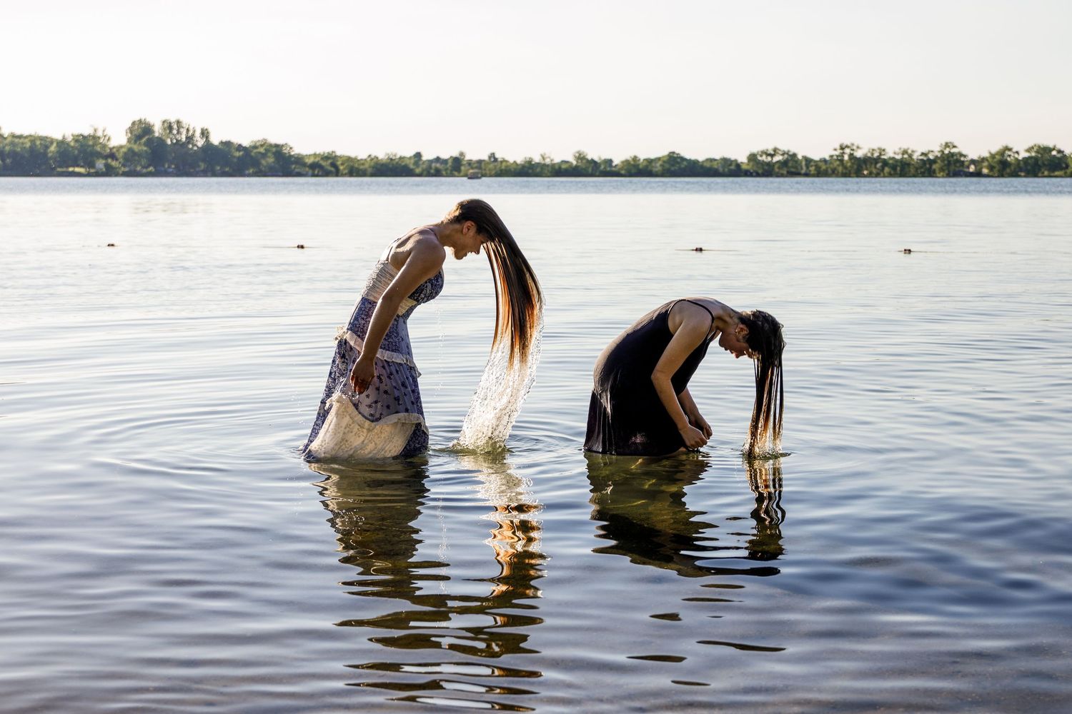 Two women standing in a lake bending forward letting their wet hair hang down over the water.