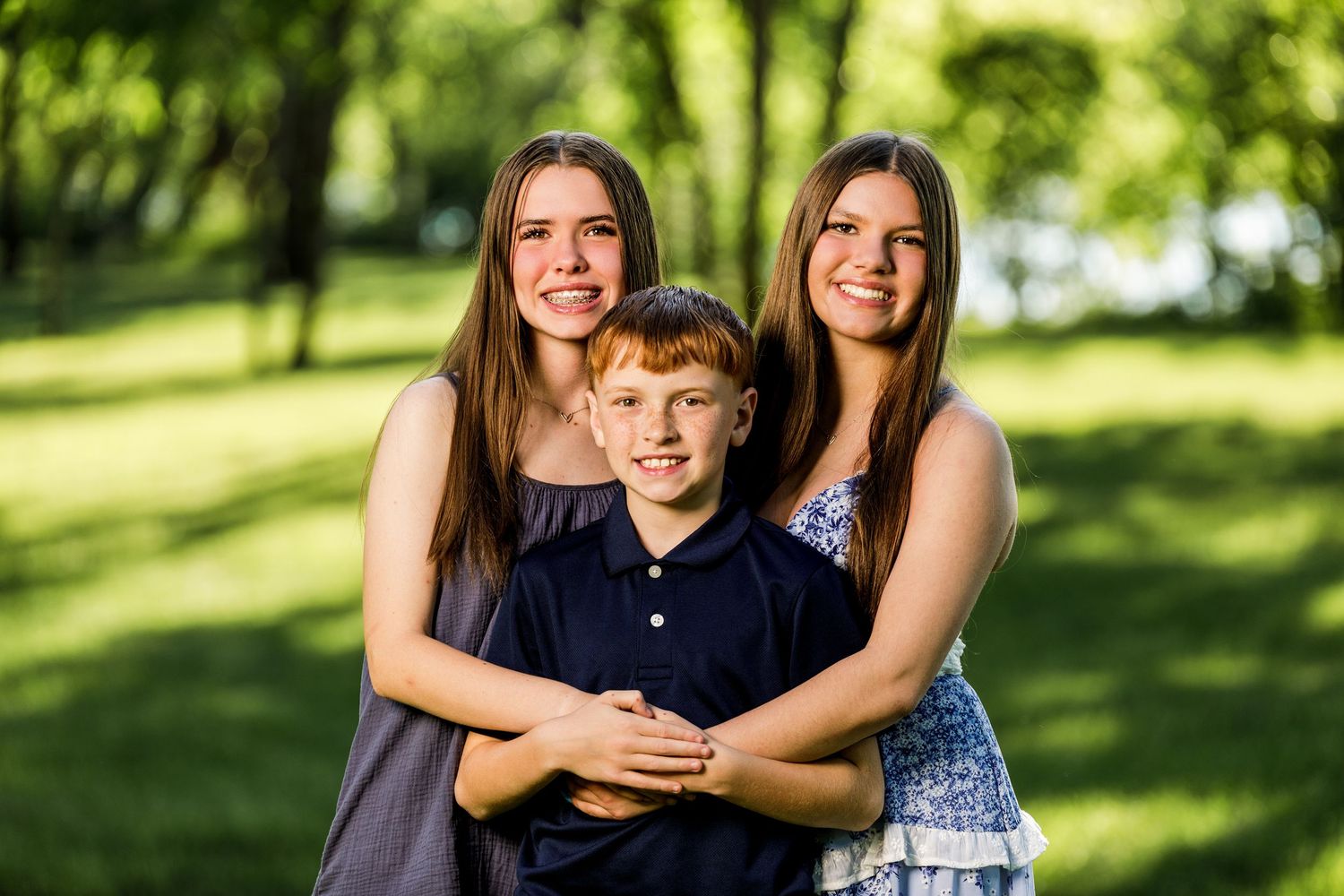 Two teenage girls hugging a young red-haired boy while posing together in a lush green park.