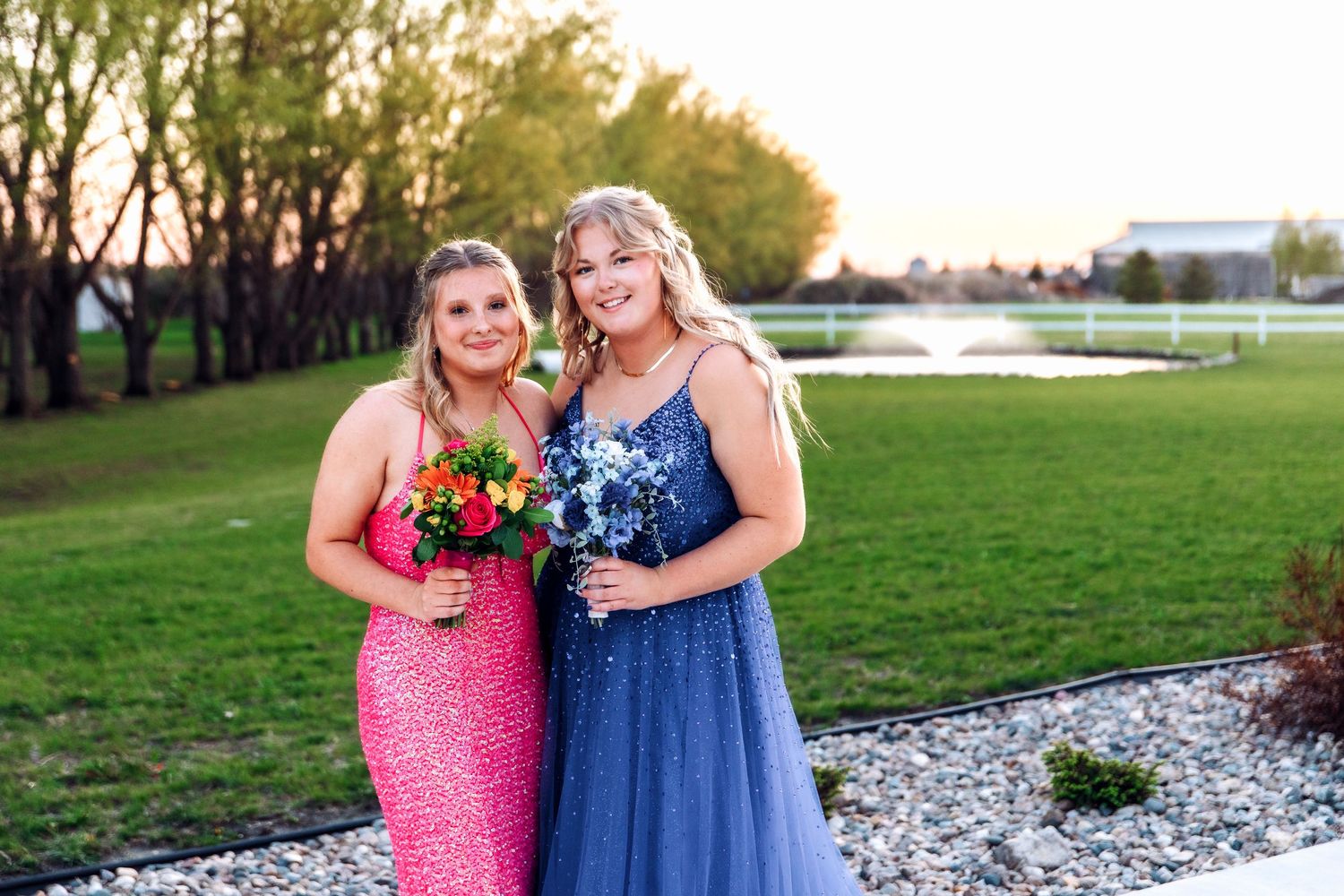 Two people in pink and blue dresses standing together outdoors in a garden setting.
