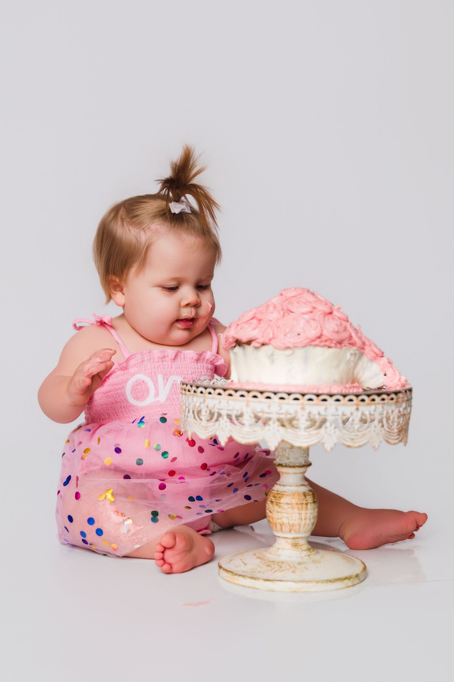 Baby in pink polka dot outfit exploring cake on decorative white stand during photo session.