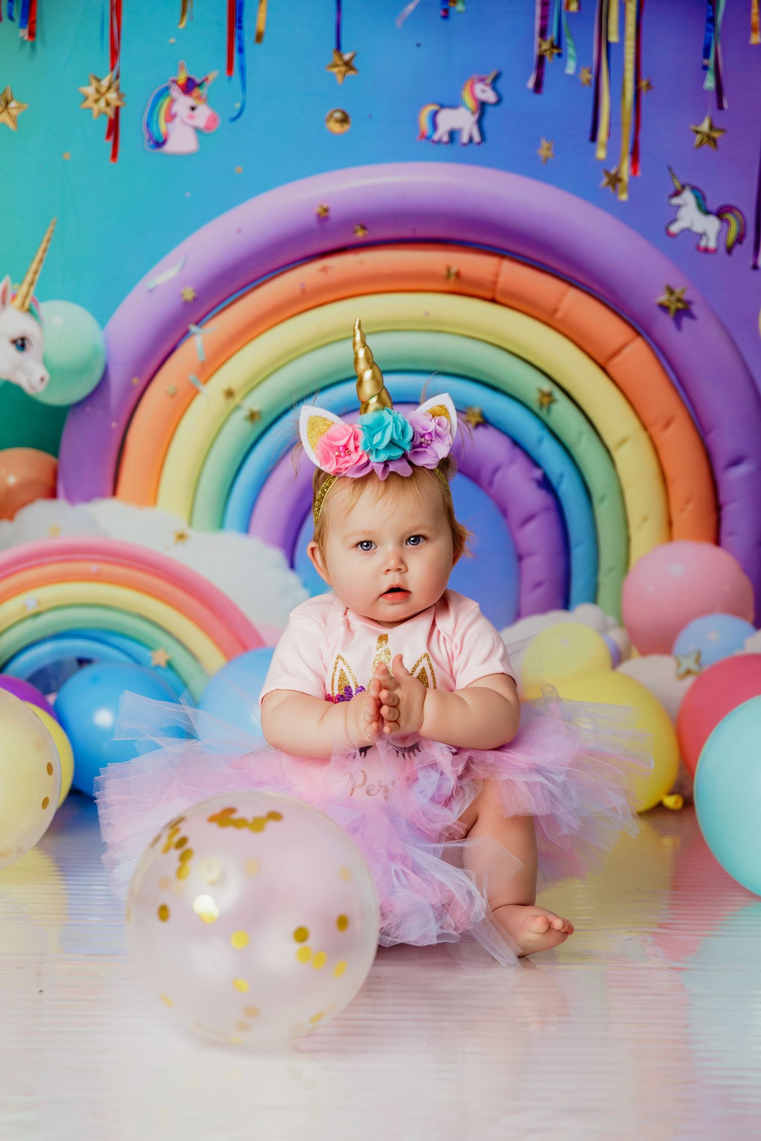 Toddler girl in pink tutu unicorn headband sits among pastel balloons at birthday party.