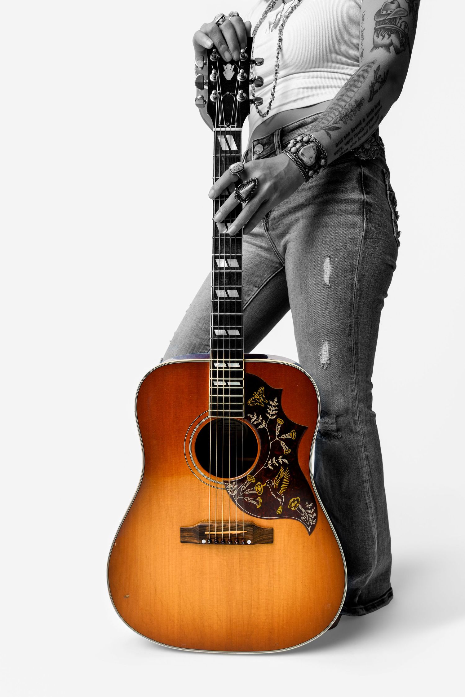 Tattooed musician holds vintage sunburst acoustic guitar in selective color photo against white background.