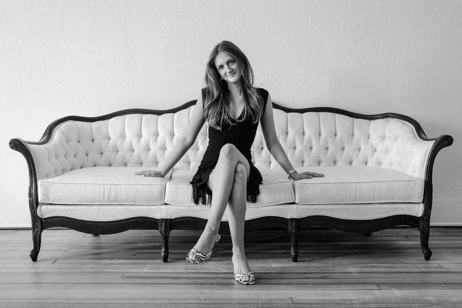 Black and white photo of a young woman in a black dress sitting gracefully on a tufted vintage sofa.