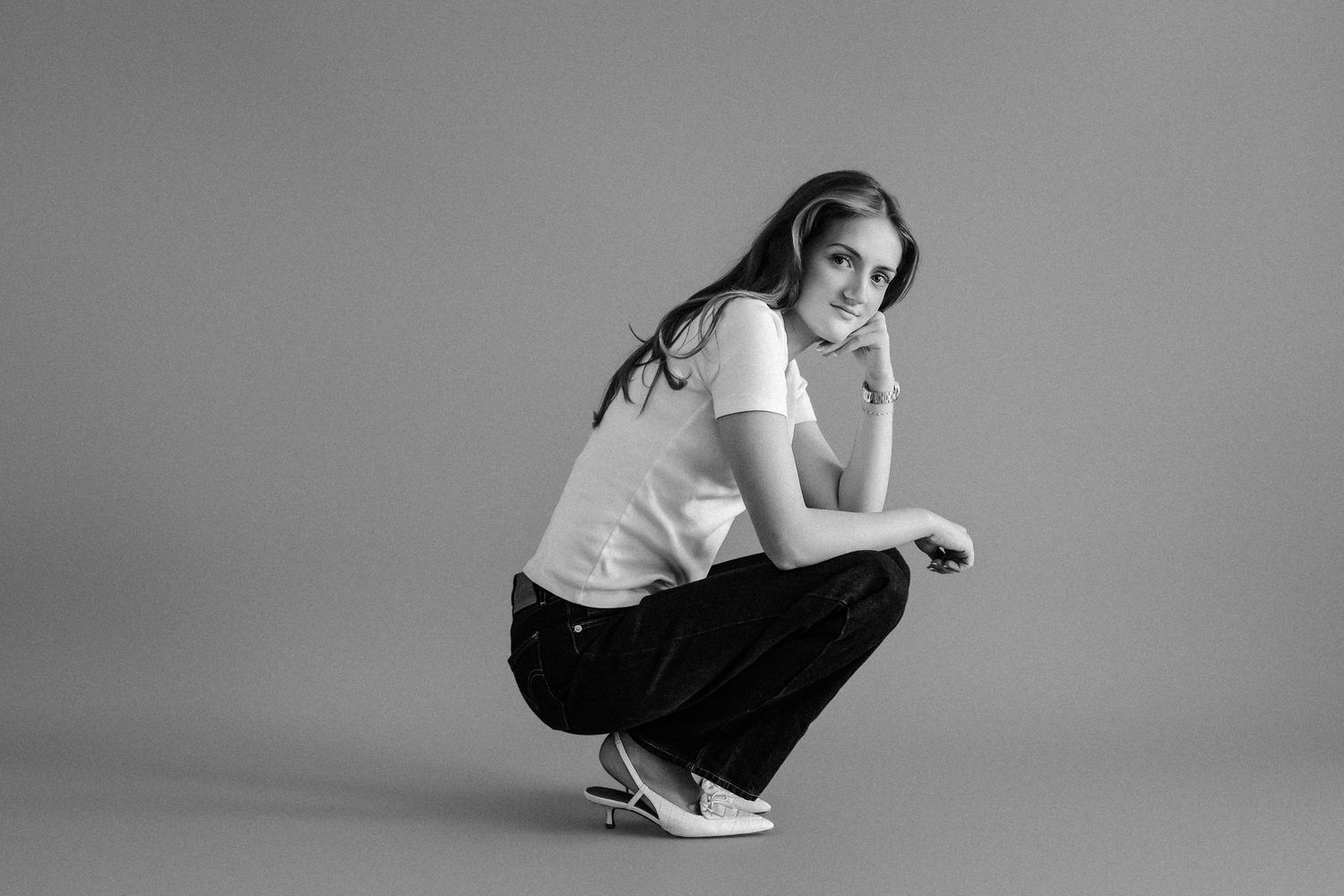 Black and white photo of a young woman crouching in white tee and dark pants against a gray backdrop.