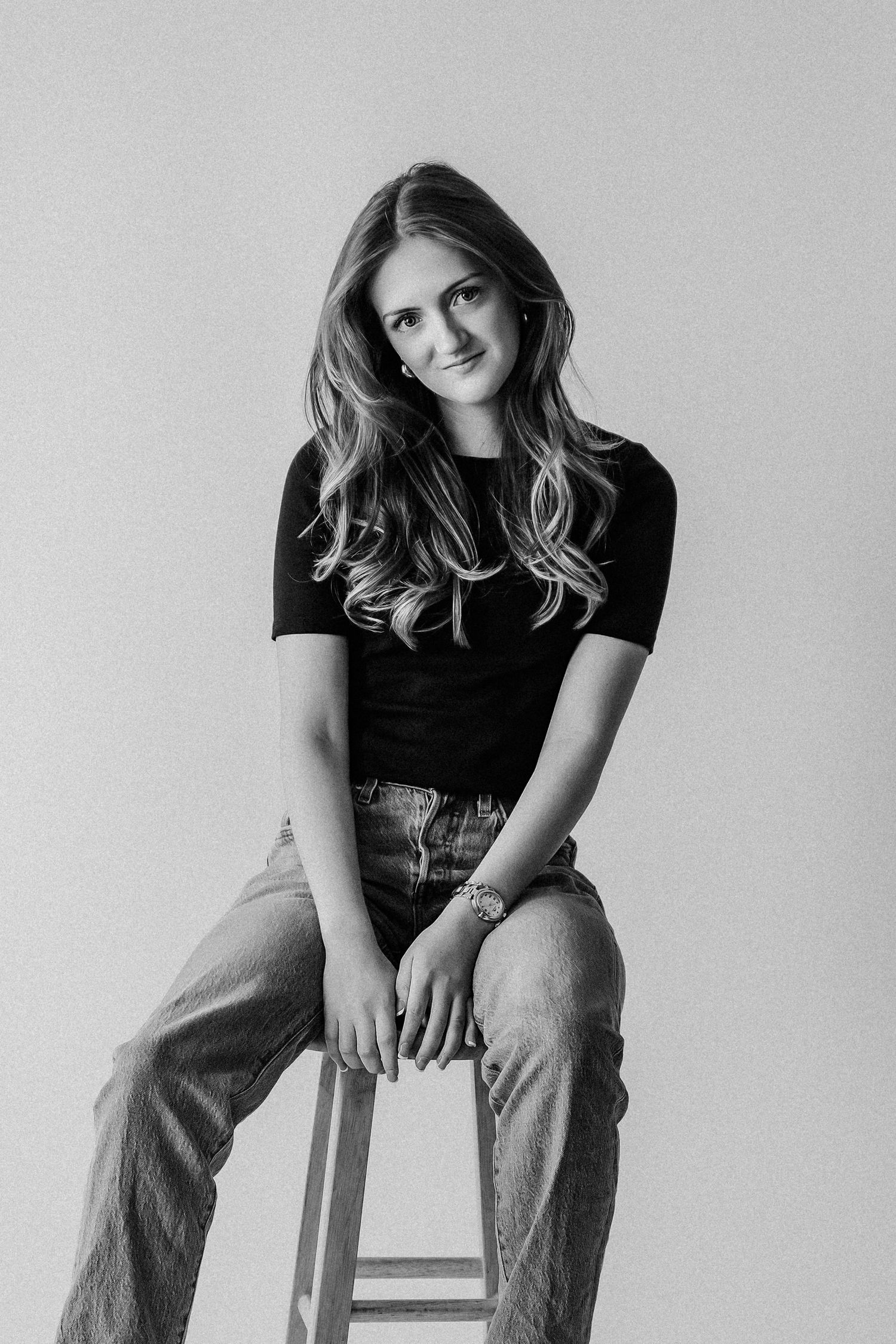 Black and white portrait of smiling young woman in black top and jeans seated on a wooden stool.