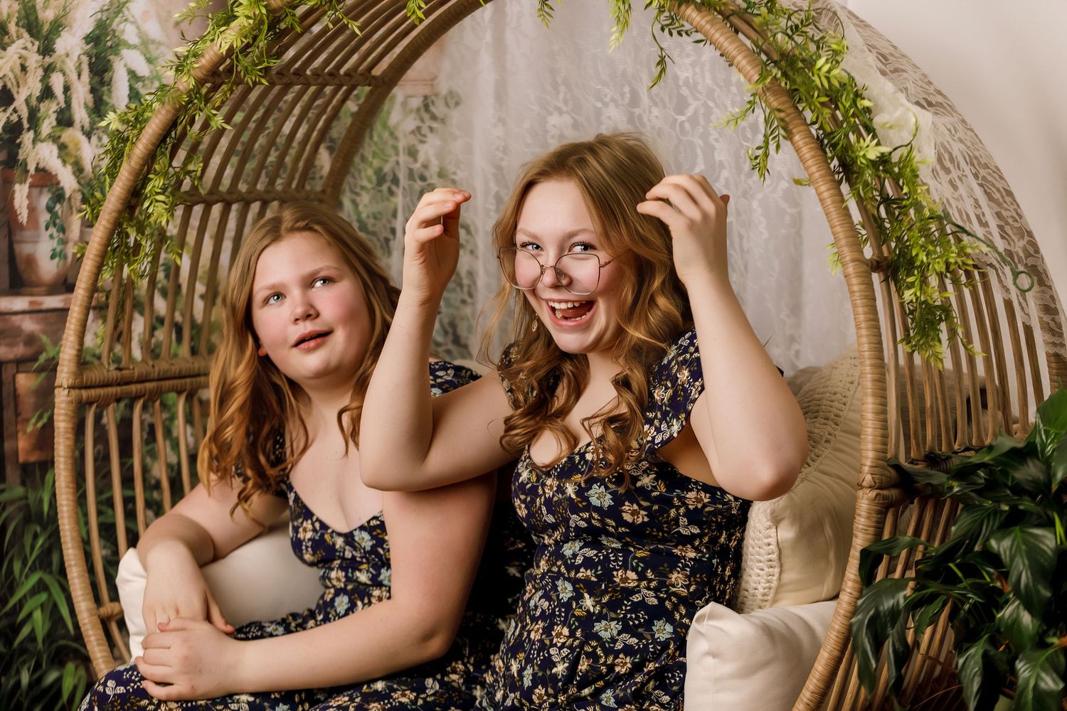 Two young women in floral dresses sitting in a large rattan swing chair surrounded by greenery.