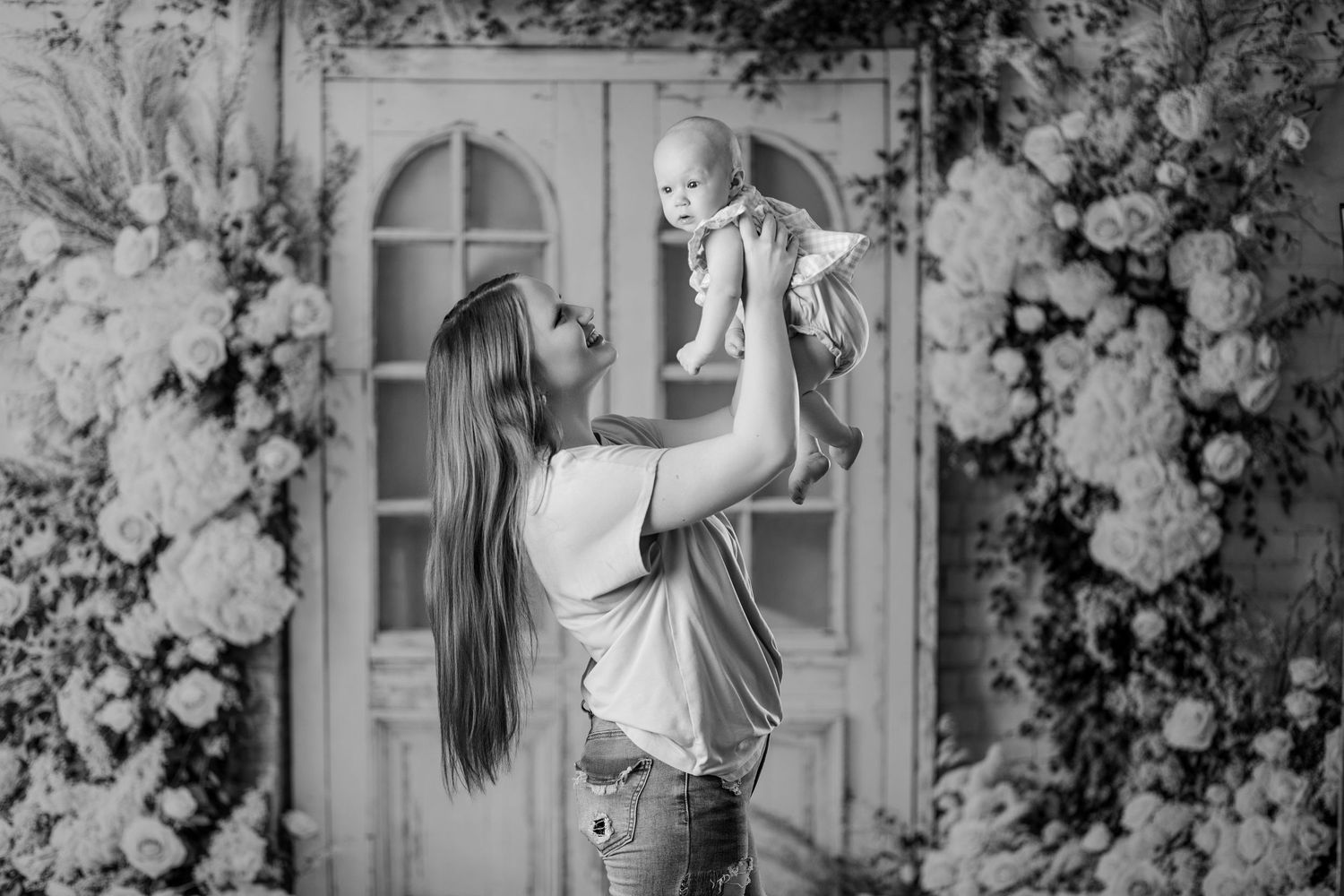 Black and white photo of a mother lifting her baby in the air surrounded by floral arrangements.