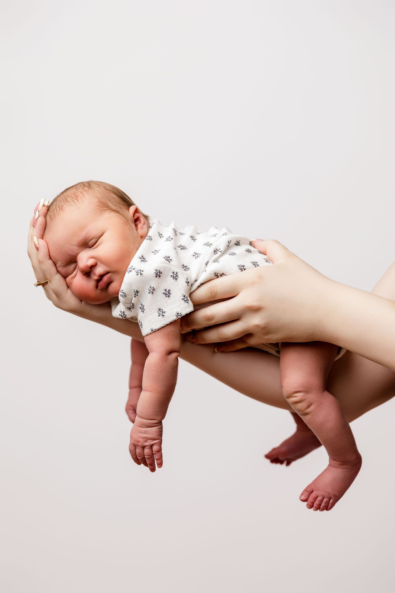 Newborn baby in white onesie held securely in adult hands against a light gray background.