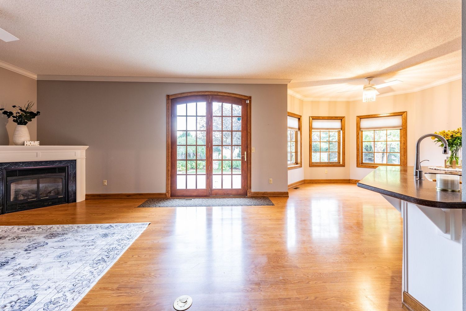 Bright open-concept living room with hardwood floors, fireplace, French doors, and large windows letting in natural light.