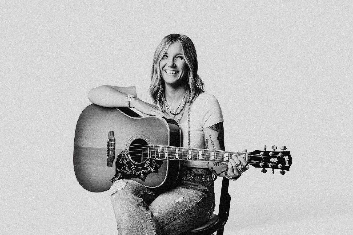 Woman smiling and playing acoustic guitar, black and white portrait photo, casual style with jeans and white t-shirt.