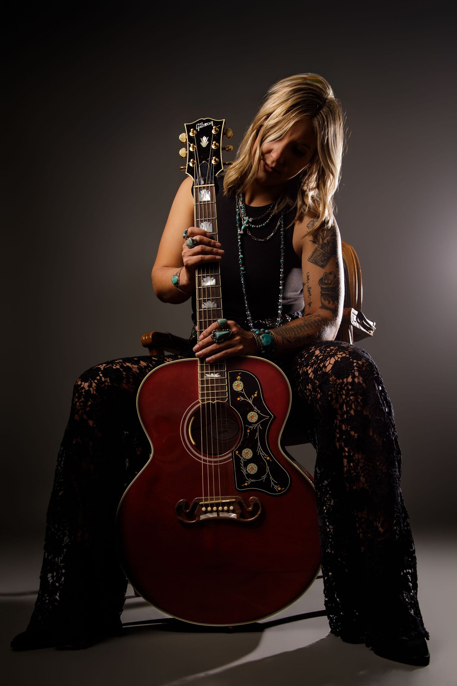 Tattooed female musician in black lace pants holding red Gibson acoustic guitar in dramatic lighting.