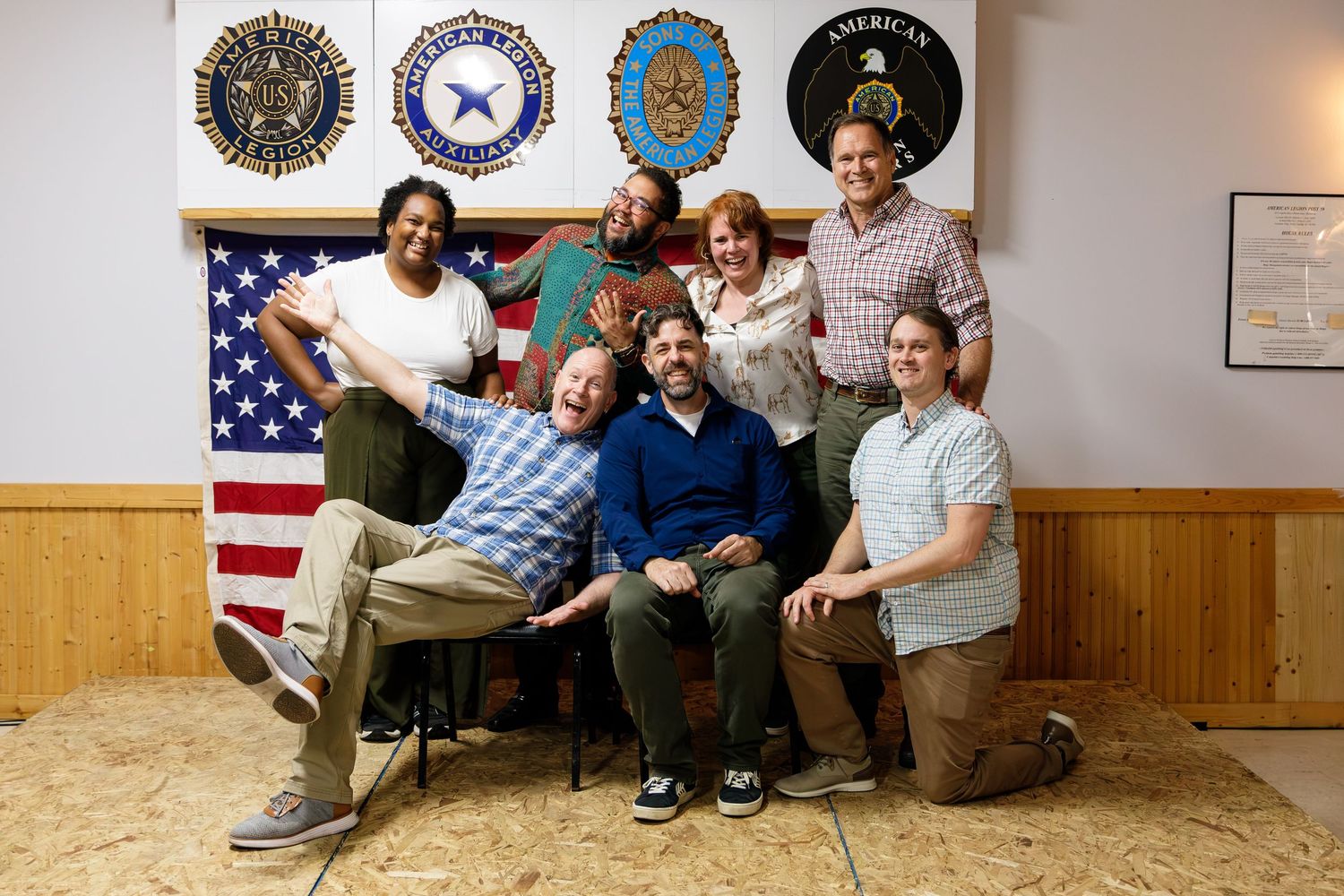 Group of seven smiling people posing together in front of an American flag and military insignia banners.