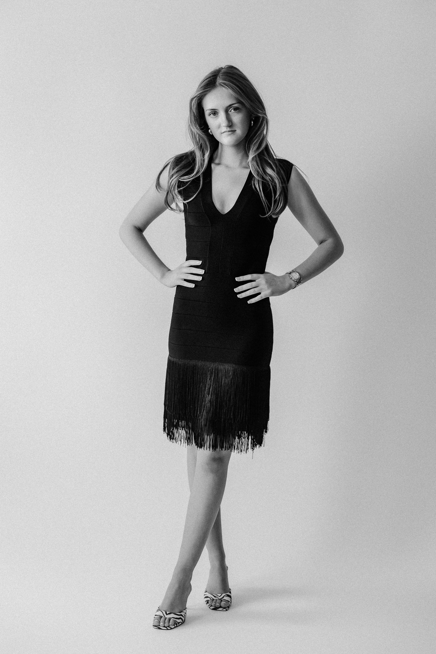 Young woman in black fringed cocktail dress and heels posing with hands on hips, black and white photo.