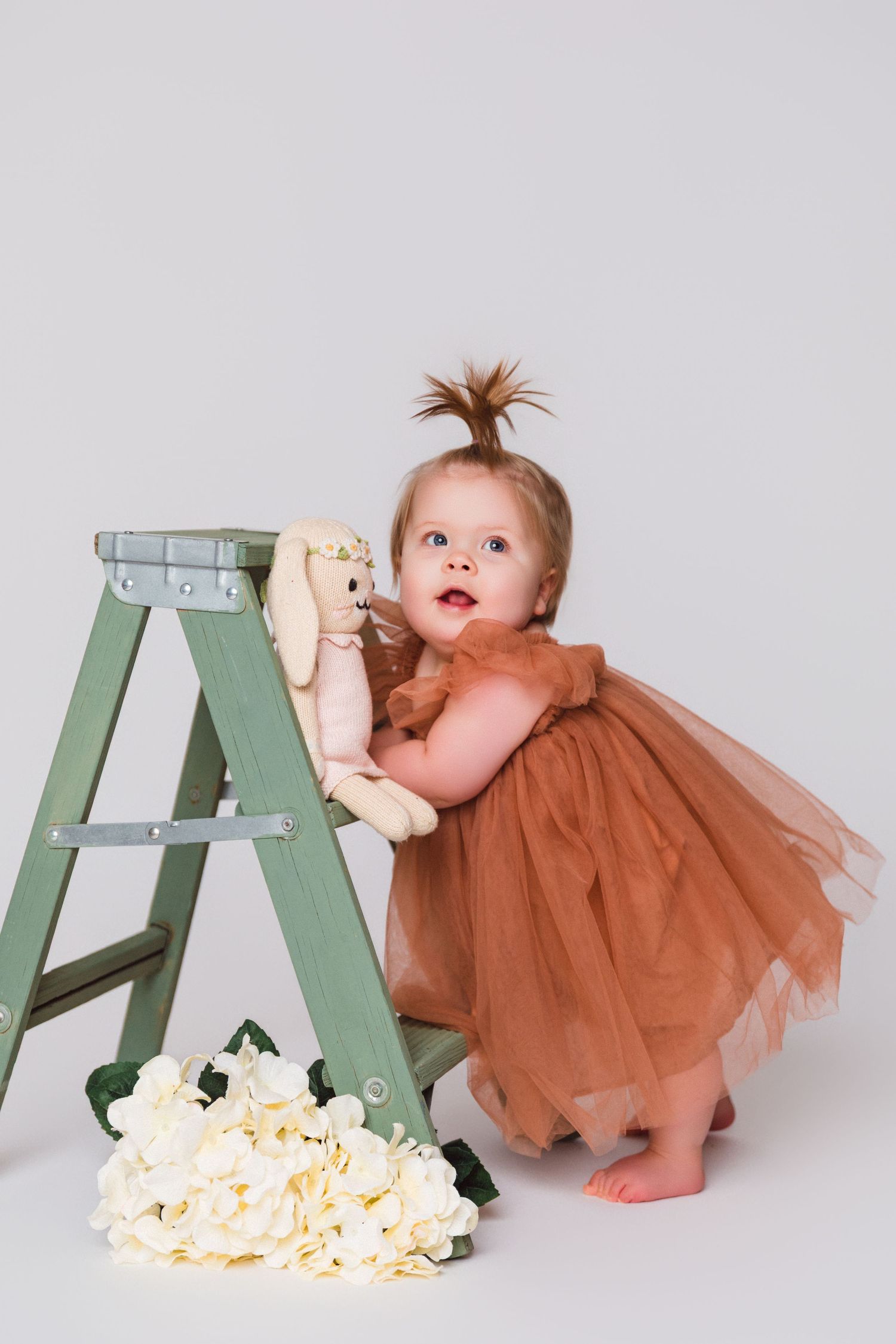 Baby in brown tulle dress playing with green ladder and scattered popcorn on floor.