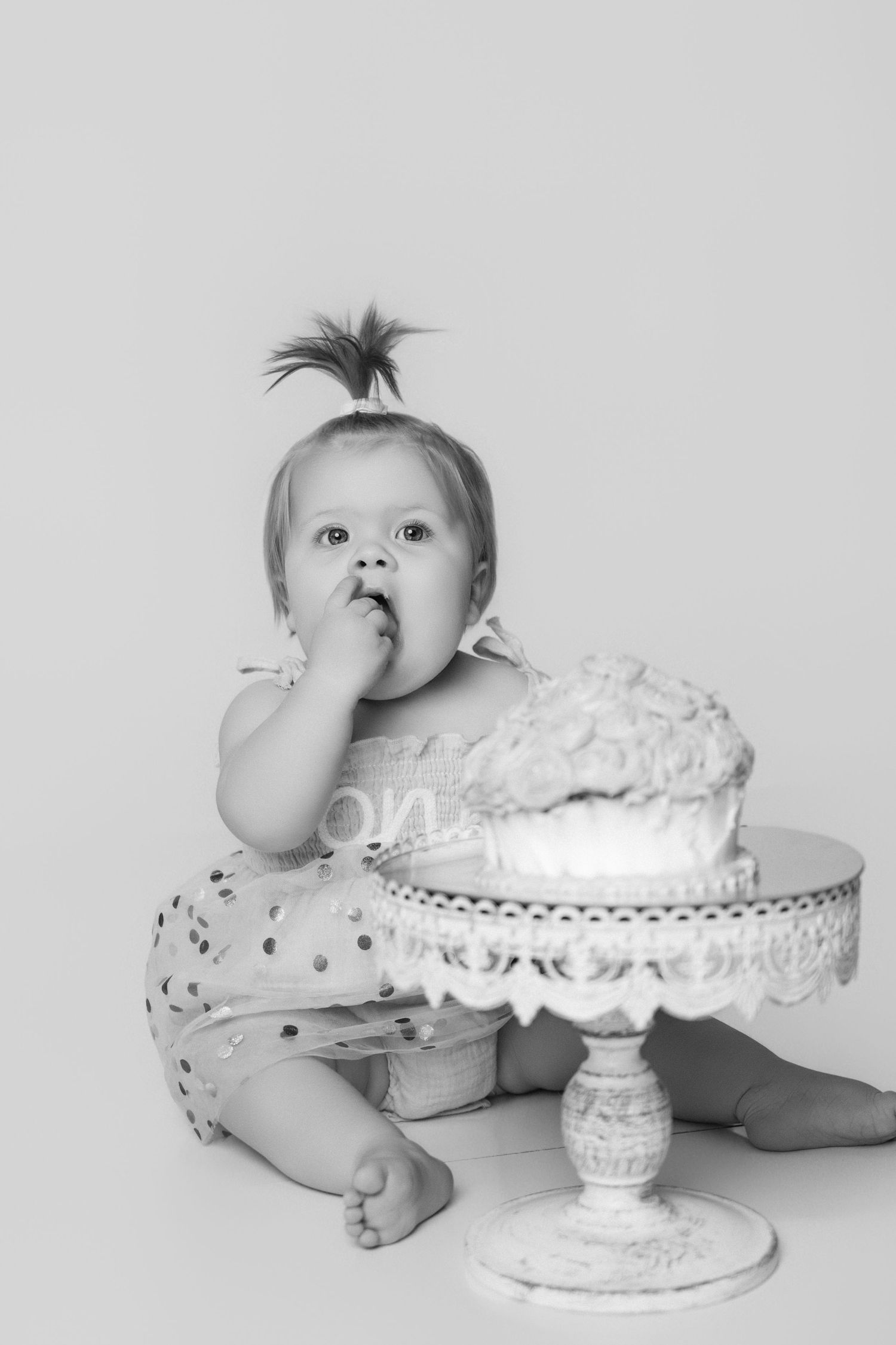 Black and white photo of baby with spiky hair reaching for cake on decorative cake stand.