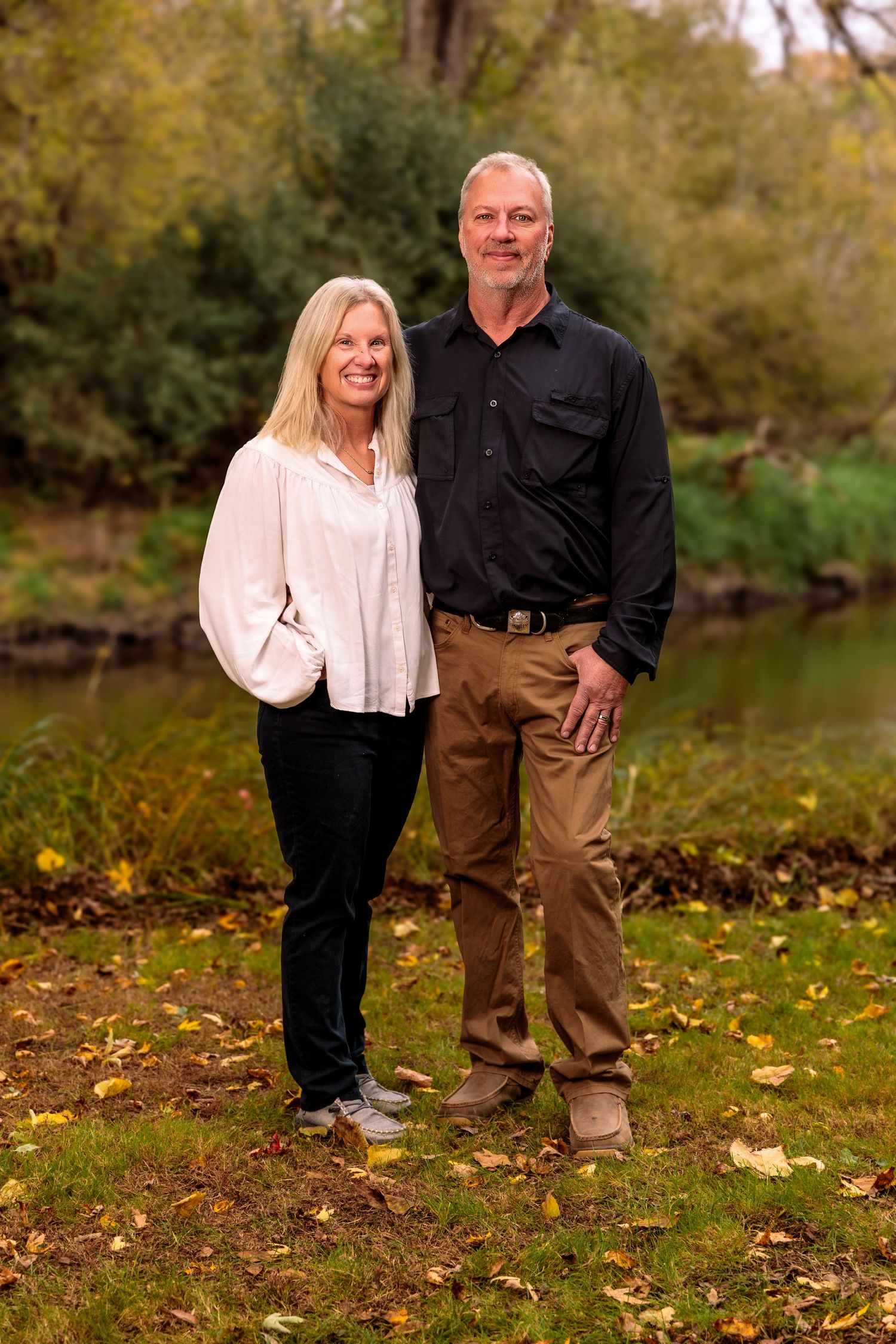 Couple posing for photos by a pond with autumn leaves on the ground.